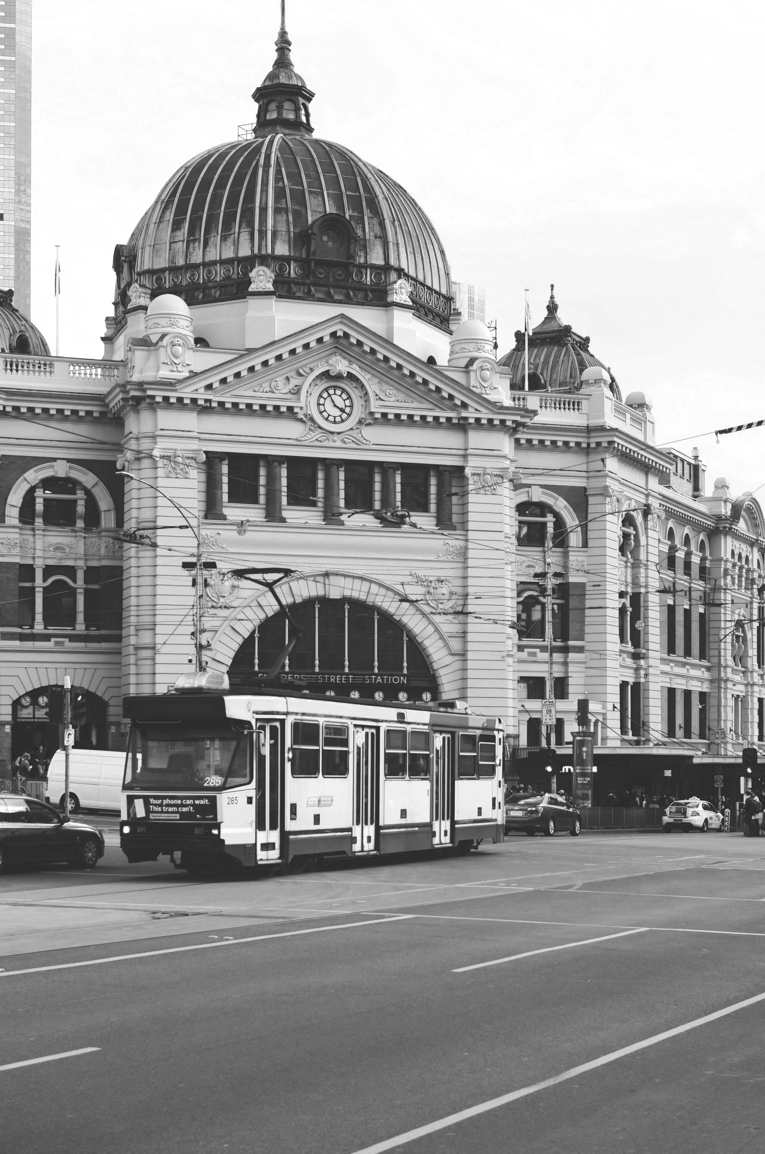A city street scene in black and white, with a bus in the foreground, taxis and cars, and an ornate historic building with a large clock and domed roof in the background.
