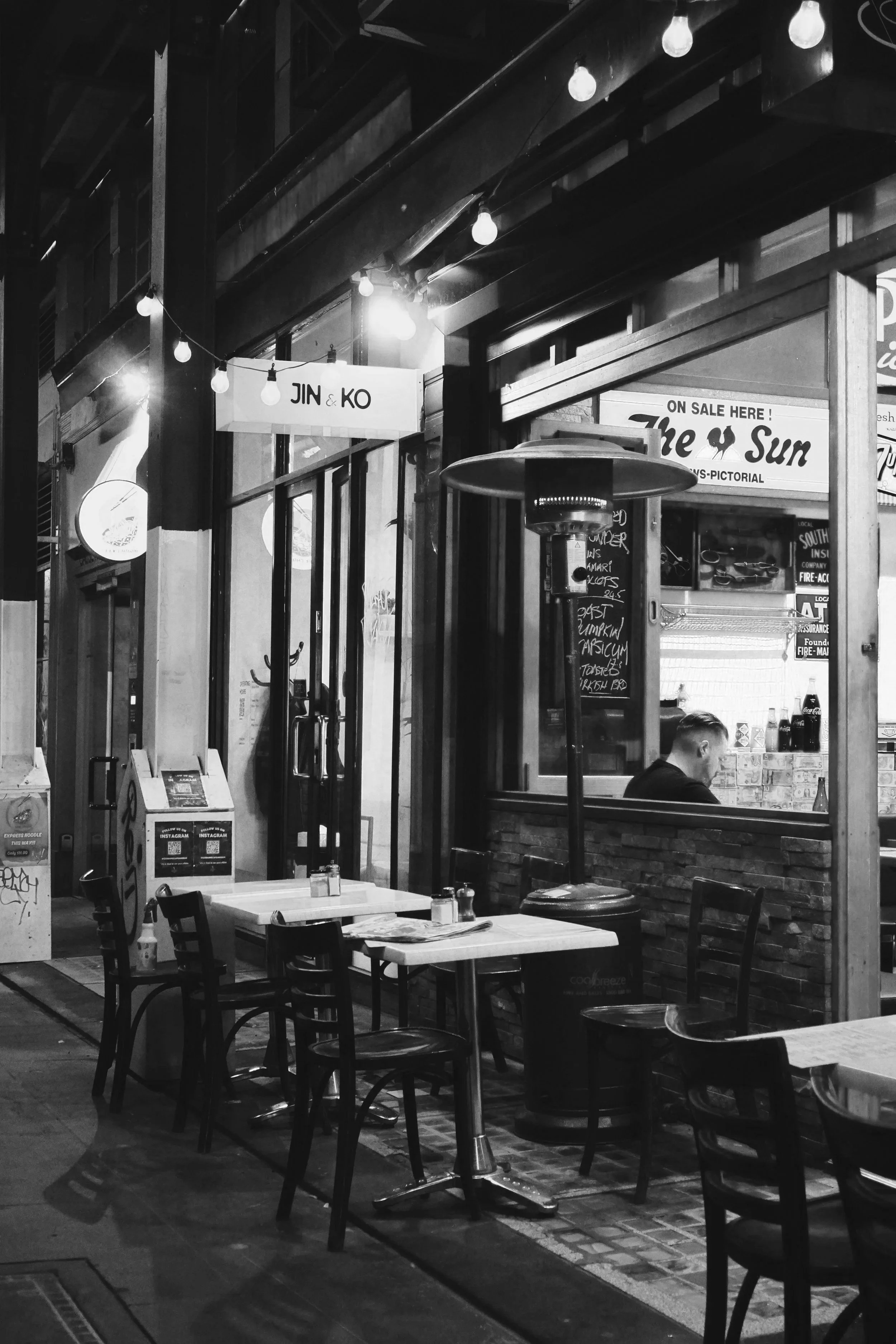 A black-and-white photo of a restaurant or café interior at night with empty tables and chairs, a heater, and illuminated signs for Jin & Ko and The Sun, with some people seated inside.