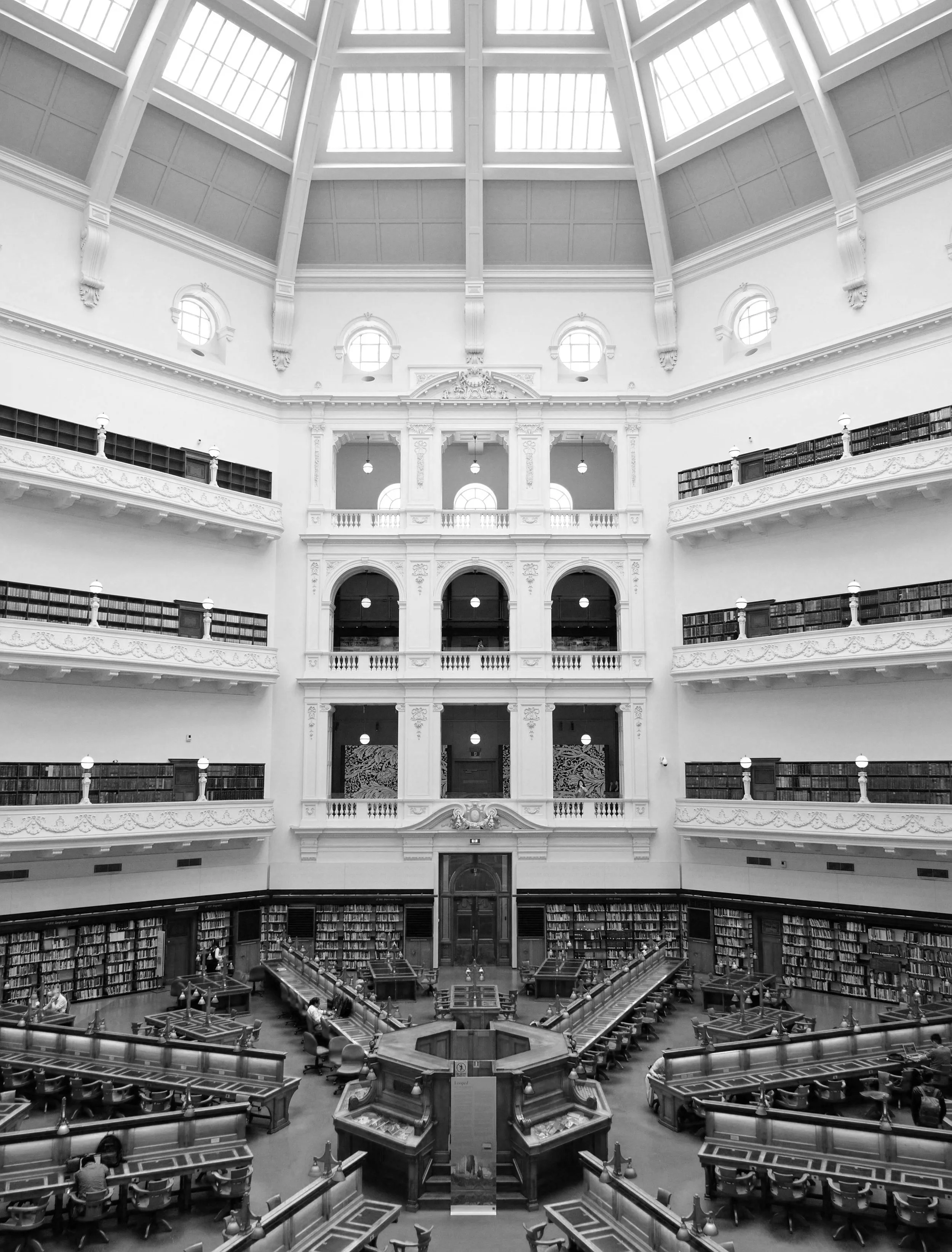 A large, ornate library hall with multiple balconies, tall windows, and a high, domed ceiling with skylights. Inside, there are tables and chairs arranged for reading and study, and bookshelves lining the walls.