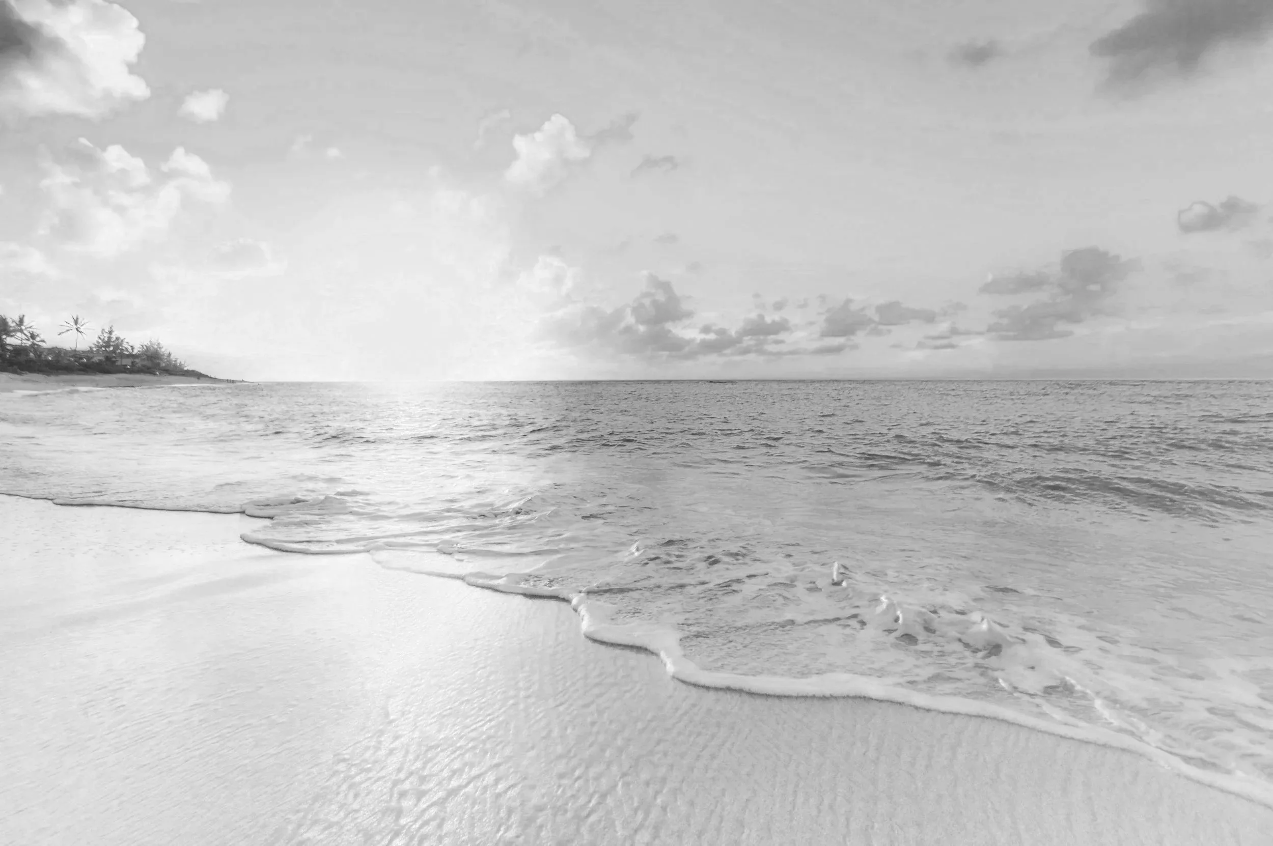 Black and white photo of a calm beach with gentle waves reaching the sandy shore, a few trees on the left side, and a partly cloudy sky above.