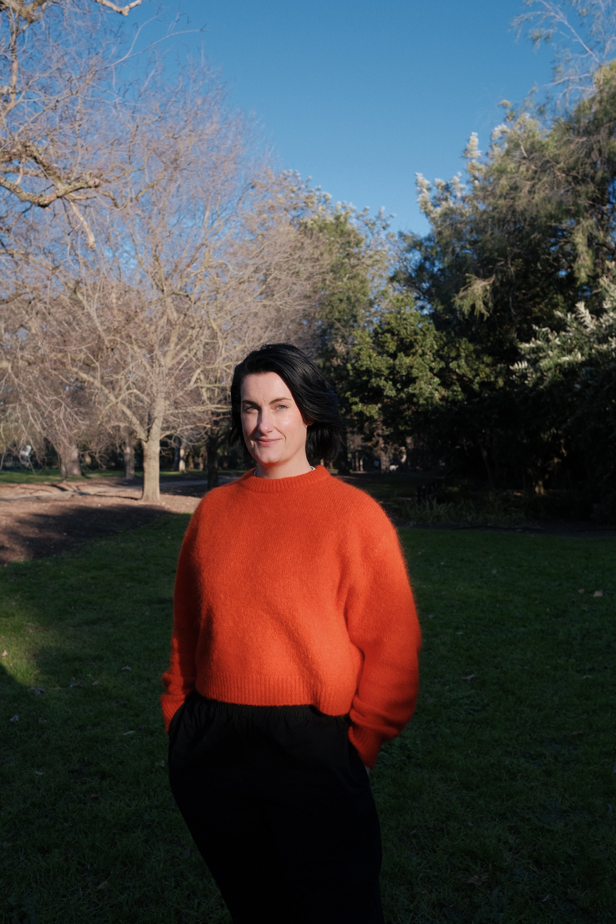 A non-binary person with short dark hair wearing a bright orange sweater and black pants stands outdoors in a park during late afternoon or early evening, with trees and a clear blue sky in the background.