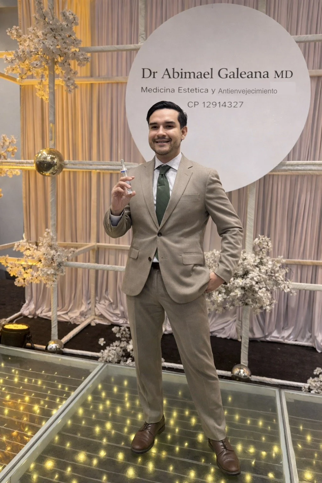 Un hombre con traje beige y corbata verde sostiene una jeringa en una celebración, con un cartel que dice 'Dr Abimael Galeana MD, Medicina Estética y Anti-envejecimiento' y decoraciones florales y de luces.