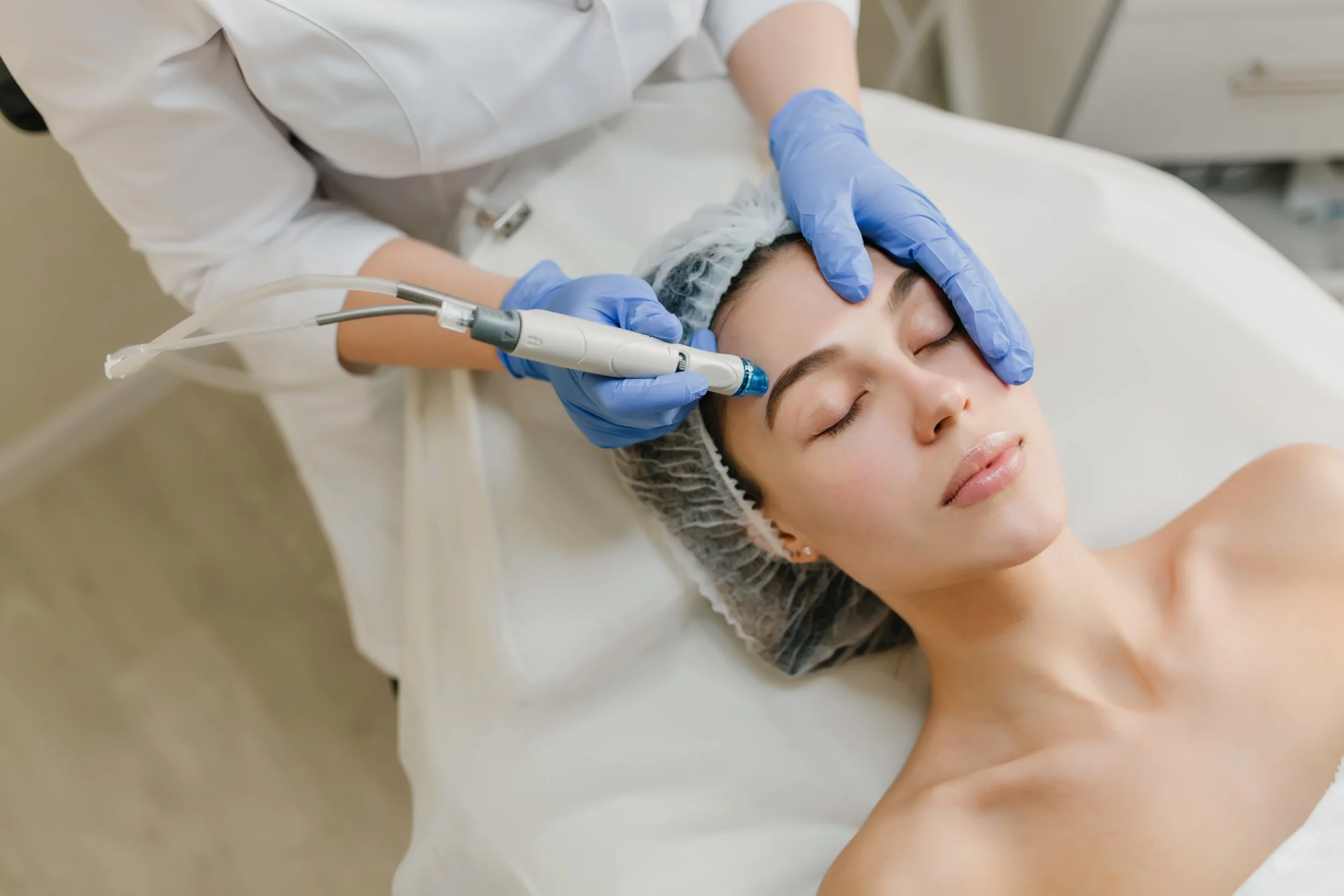 Paciente recibiendo un procedimiento estético facial de un profesional en un entorno clínico, con guantes y mascarilla, en una camilla.
