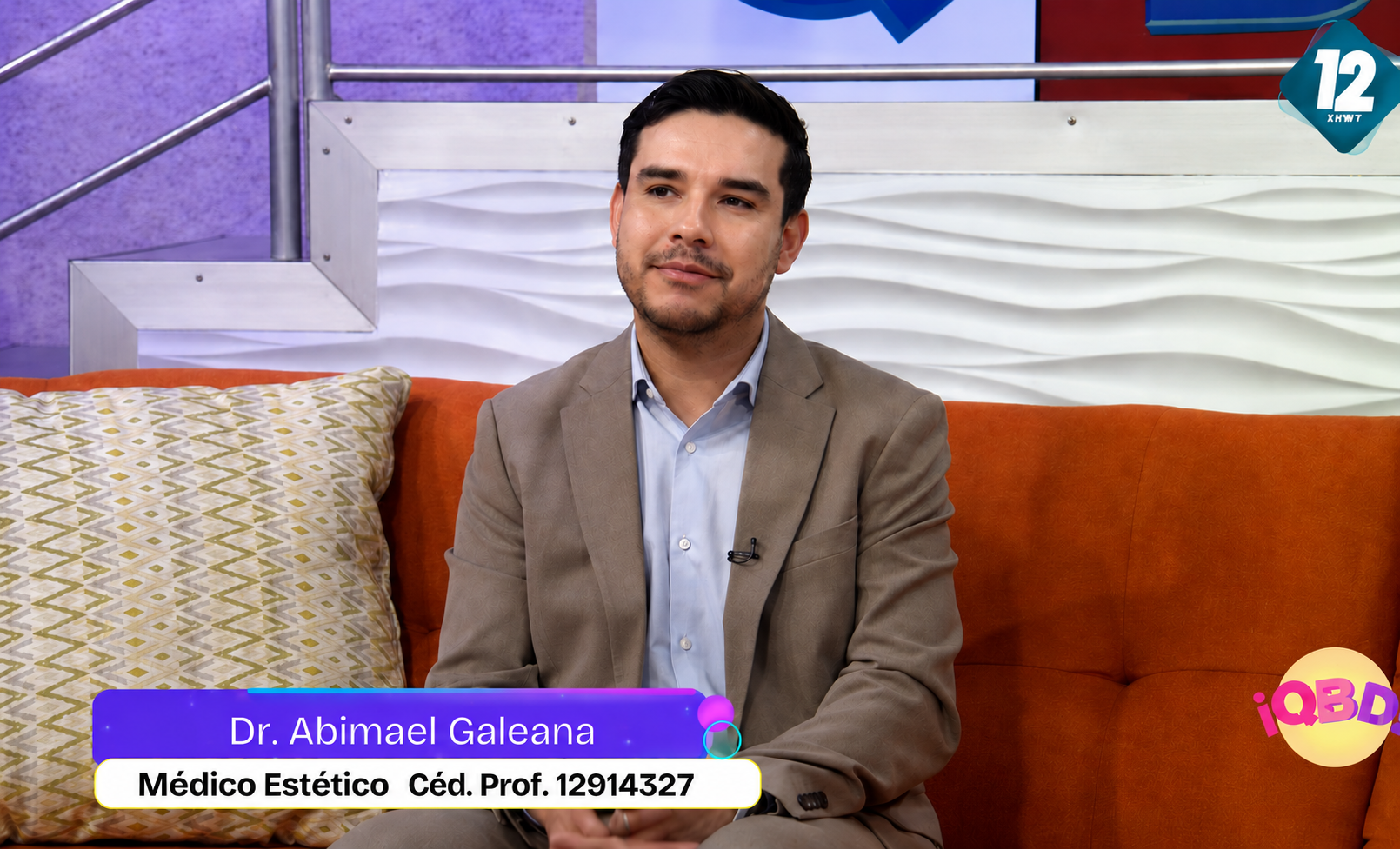 Un hombre sentado en un sofá naranja, usando un saco gris y camisa azul, en un programa de televisión, con un cartel que dice 'Dr. Abimael Galeana, Médico Estético, Céd. Prof. 12914327', en un set con paredes blancas y decoraciones gráficas.