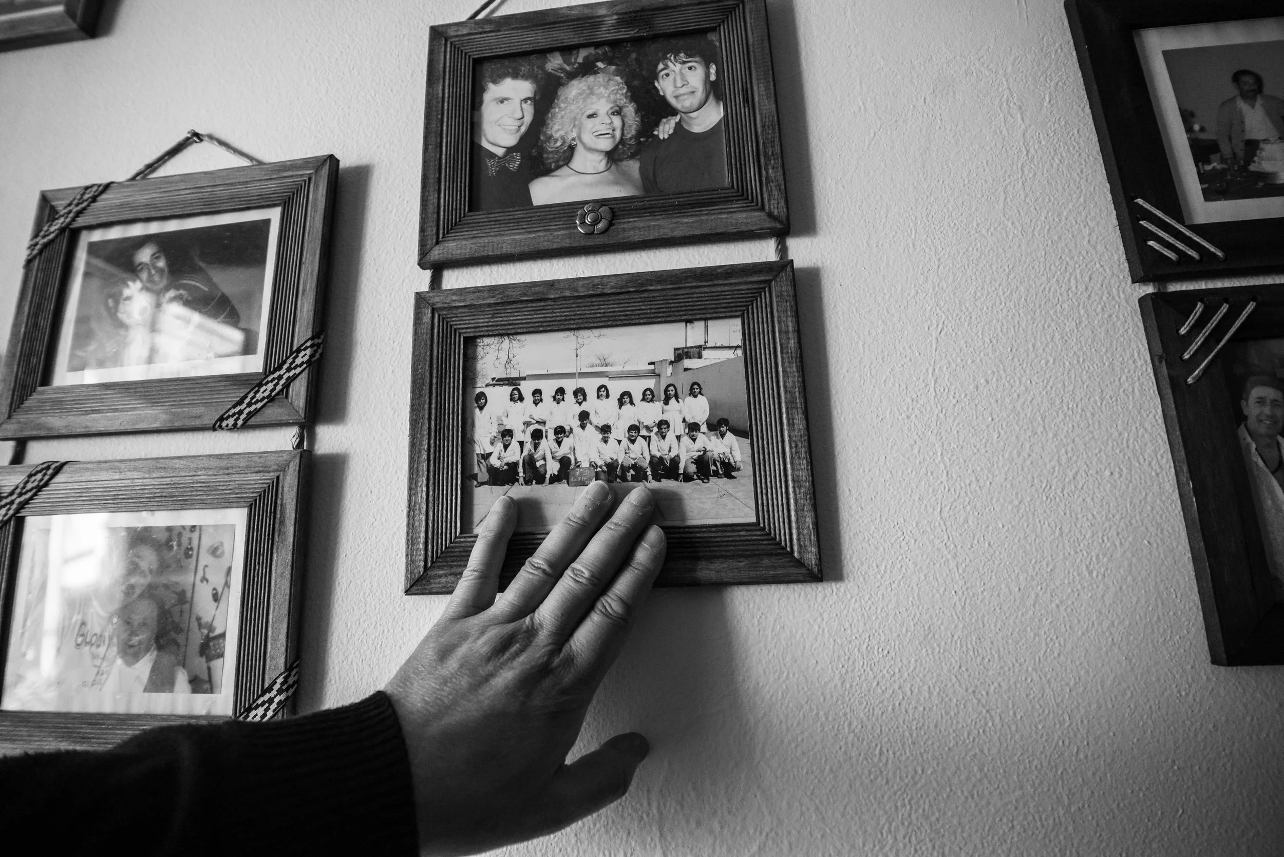 Black and white photo of a hand holding a framed photo of a group of people in jewelry and formal attire, with other framed photos hanging on a wall.