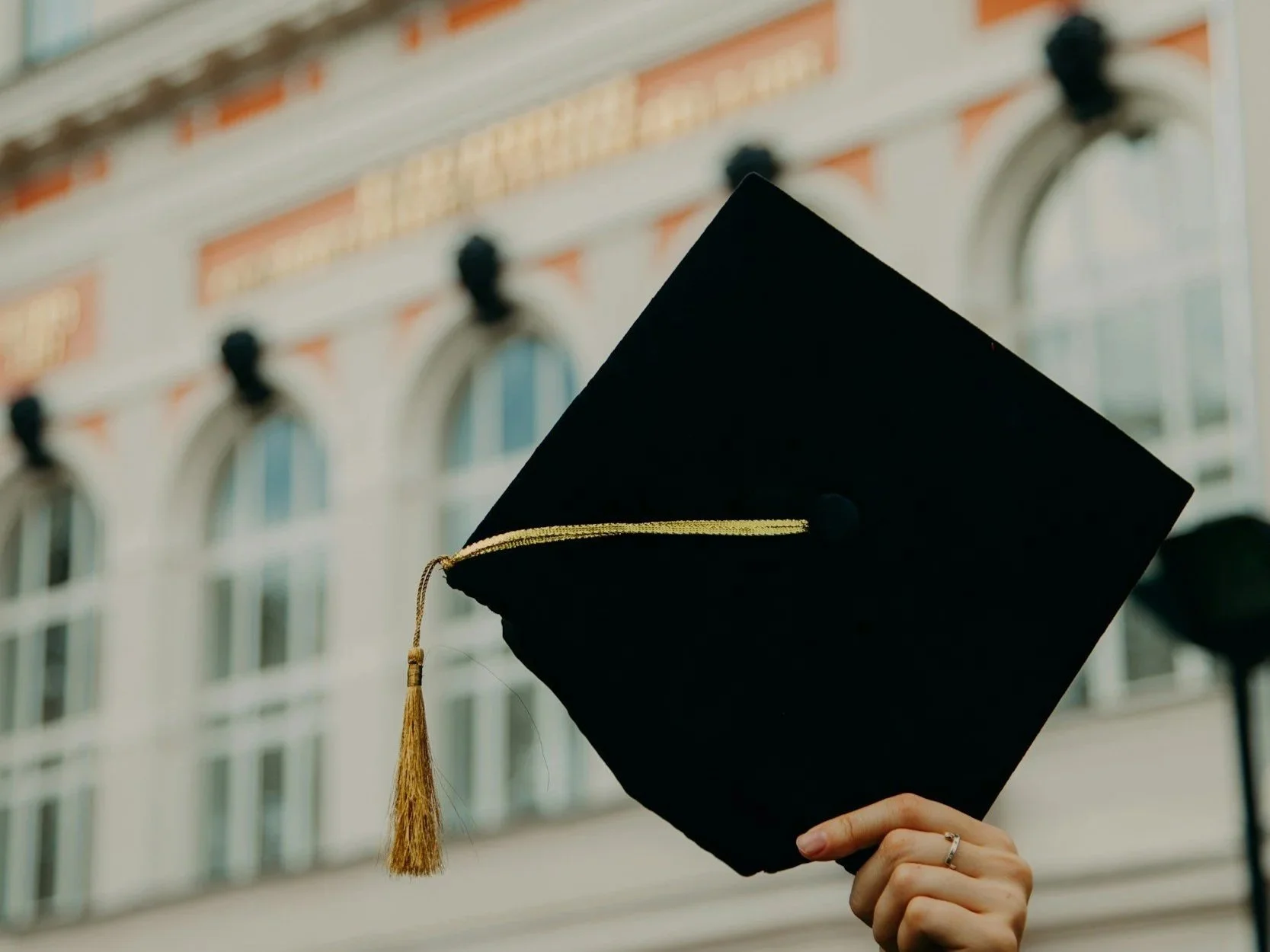 A person holding a black graduation cap with a gold tassel in front of a historic building with large windows and ornate architectural details.