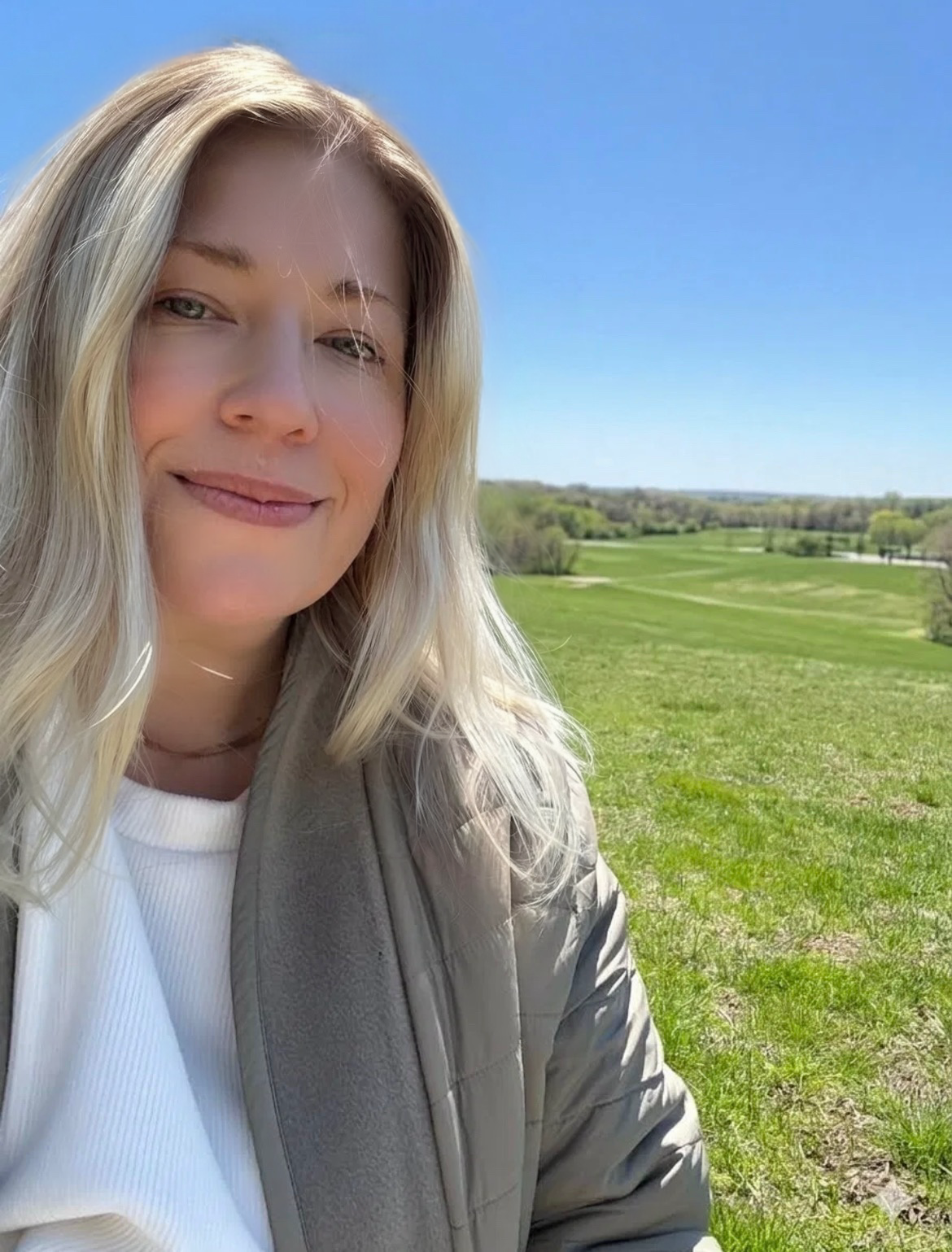 A woman with blonde hair smiling outdoors on a sunny day, with a green landscape and blue sky in the background.