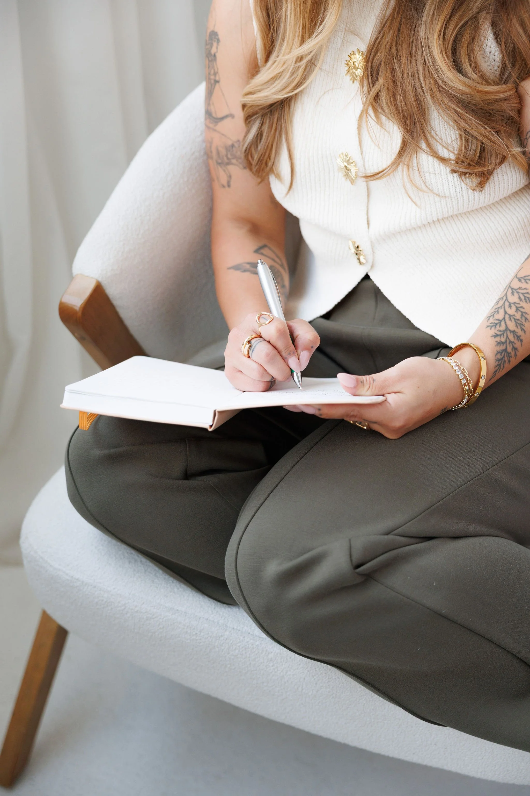 A woman is sitting on a white chair, writing in a notebook with a silver pen. She is wearing a cream-colored sleeveless top with gold floral embellishments and dark gray pants. She has multiple tattoos on her right arm and gold jewelry, including bracelets and rings.