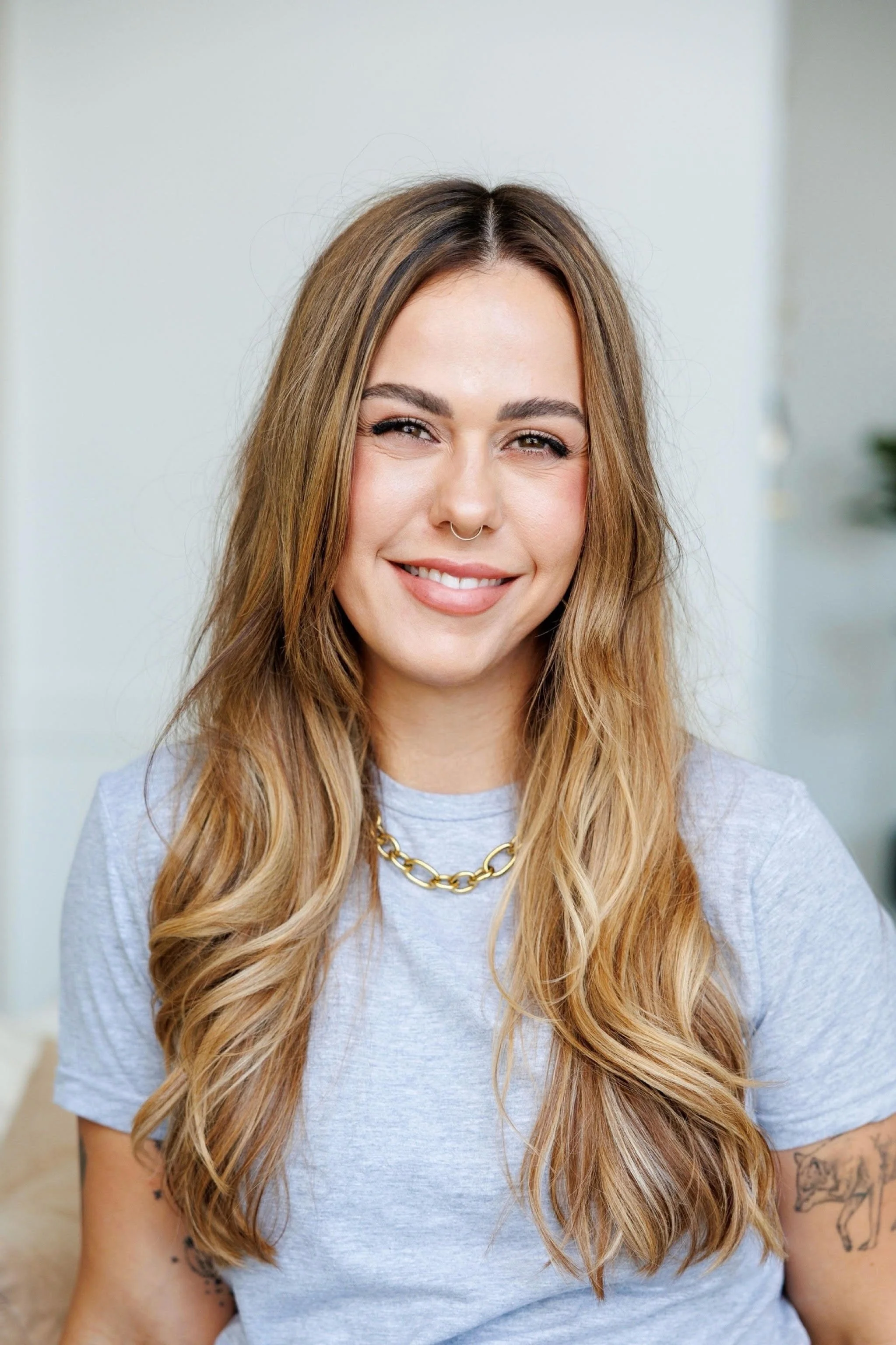 A woman with long wavy blonde hair, wearing a gray T-shirt and a gold chain necklace, smiling at the camera. She has a septum piercing and tattoos on her arms.