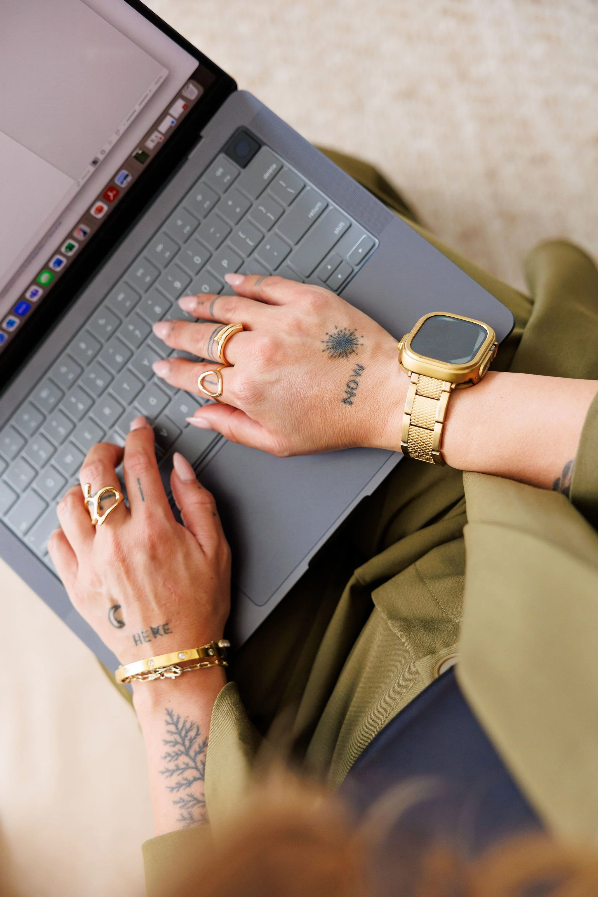 Close-up of a person's hands typing on a laptop keyboard. The person has tattoos on both hands and is wearing multiple gold rings, a gold watch, and a gold bracelet. They are wearing a green shirt or jacket.
