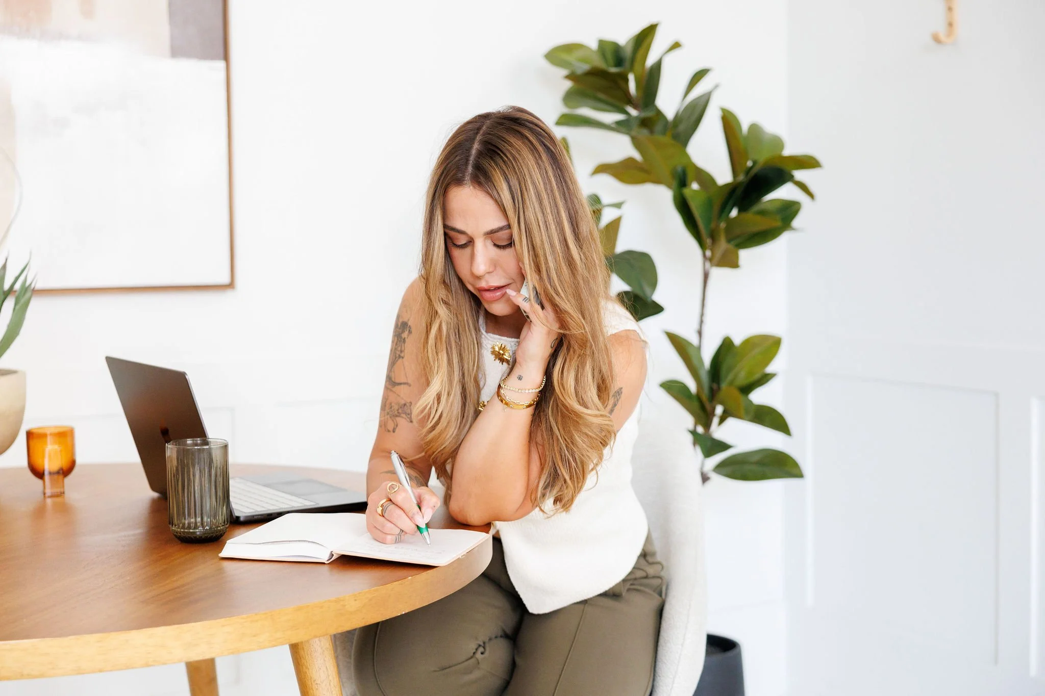 A woman with long, wavy blonde hair and tattoos on her arms is sitting at a wooden table, writing in a notebook with a pen. She is wearing a white top and beige pants, adorned with gold jewelry. On the table are a laptop, a glass of water, and an amber-colored glass. Behind her, there is a large green plant and a white wall with a picture frame.