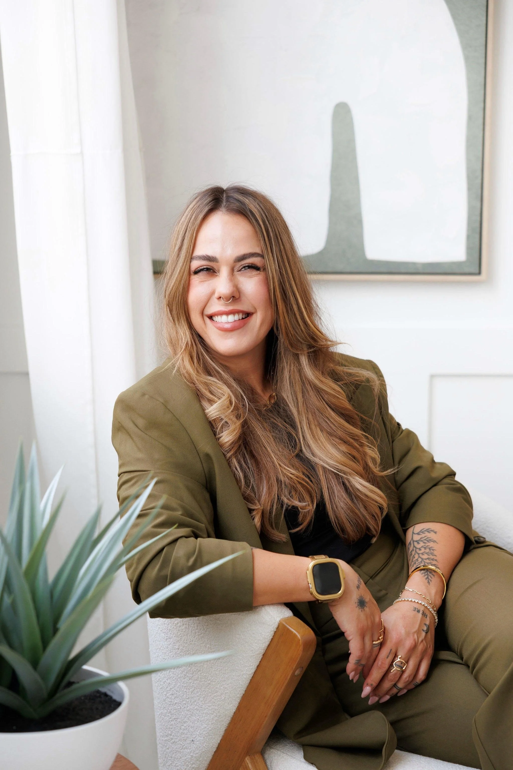 A woman with long wavy hair smiling, wearing an olive green jacket, black top, and several jewelry pieces, sitting indoors with a large framed abstract art piece behind her and a potted plant in the foreground.