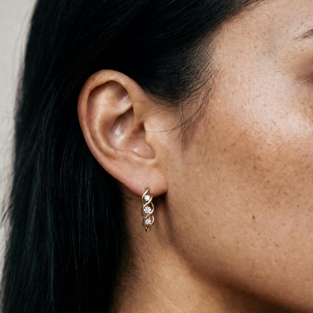 Close-up of a woman's ear with a gold earring featuring three diamonds in a twisted design. Her dark hair is tucked behind her ear, revealing skin with freckles.