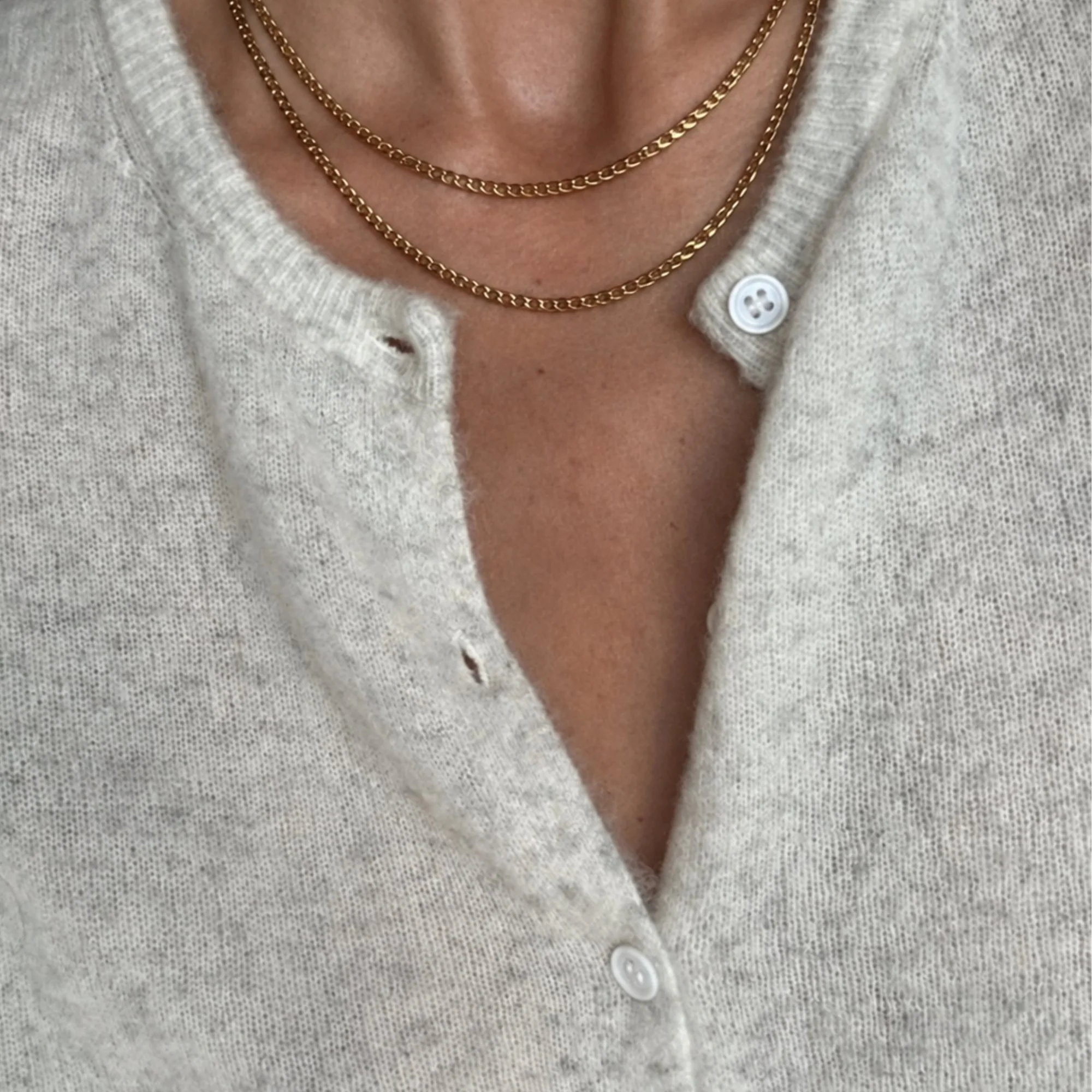 Close-up of a woman's neck wearing three gold chains, a white button-up shirt, and a beige knitted cardigan.
