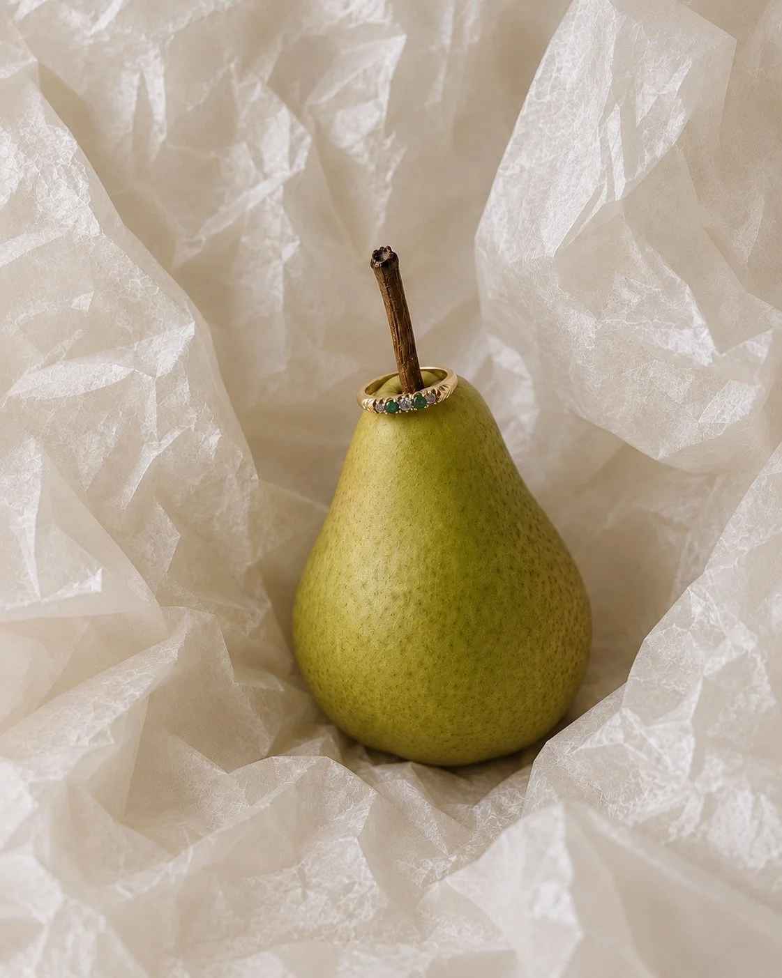 A yellow pear-shaped fruit with a ring of gold and green stones around its opening, with a ring through the stem, placed on crumpled white tissue paper.