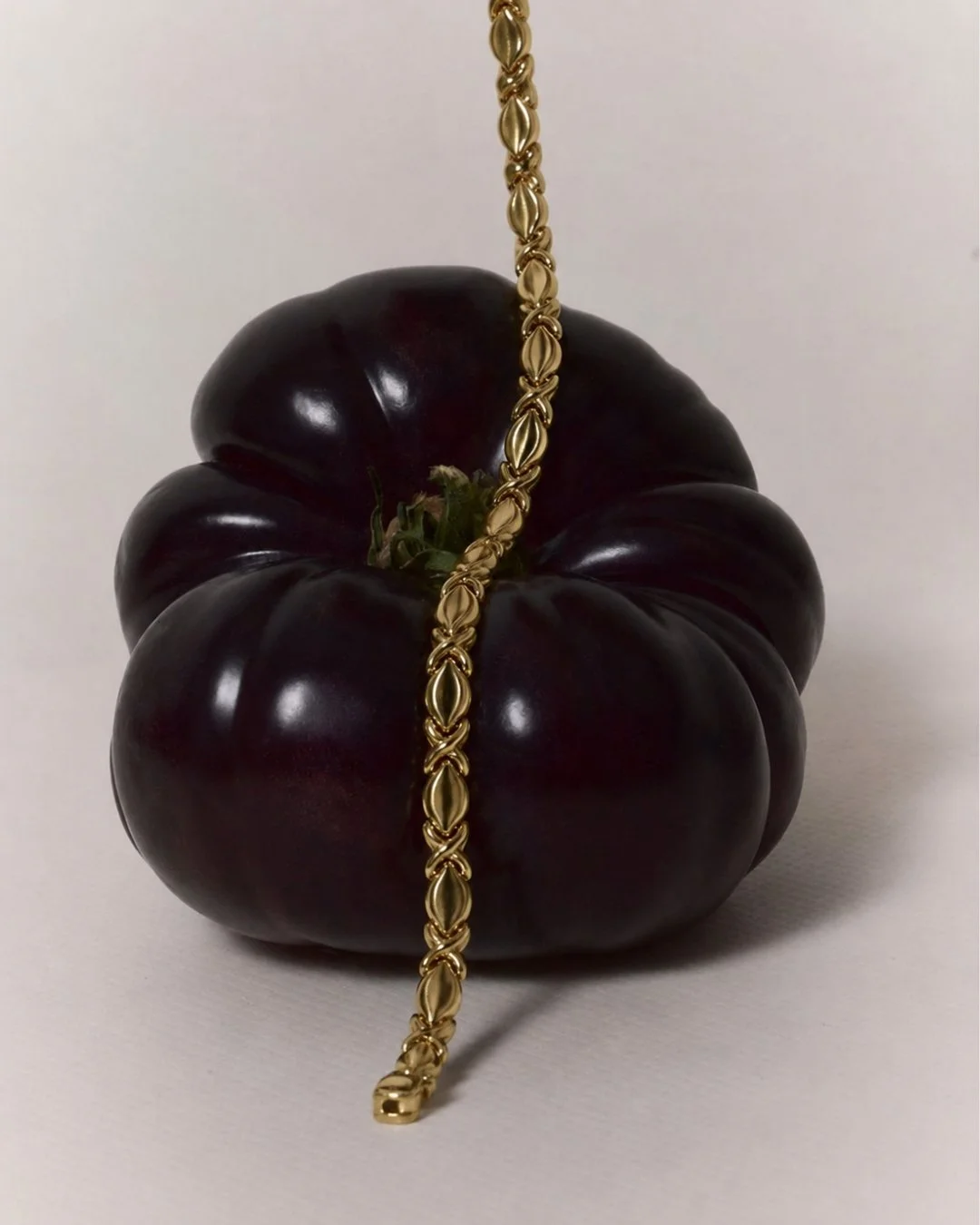 A creative still-life for Vallora Vintage: a dark purple heirloom tomato draped with a thick vintage gold chain, set against a plain light background.