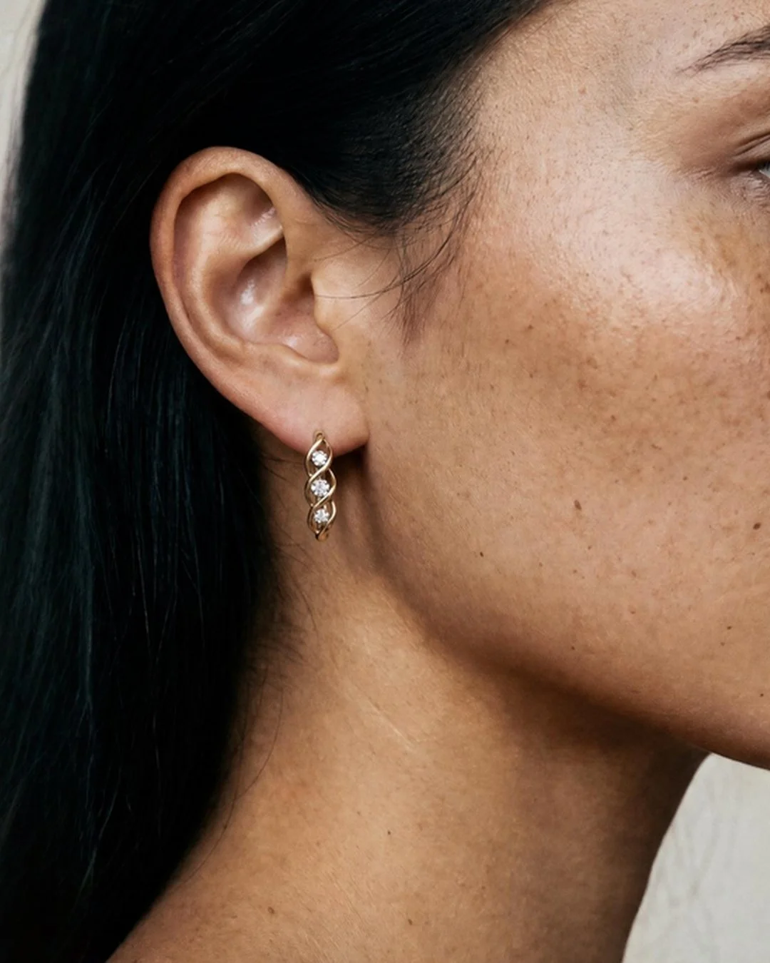 Close-up of a model wearing a gold twist huggie earring with crystal detail, shown against her ear for scale