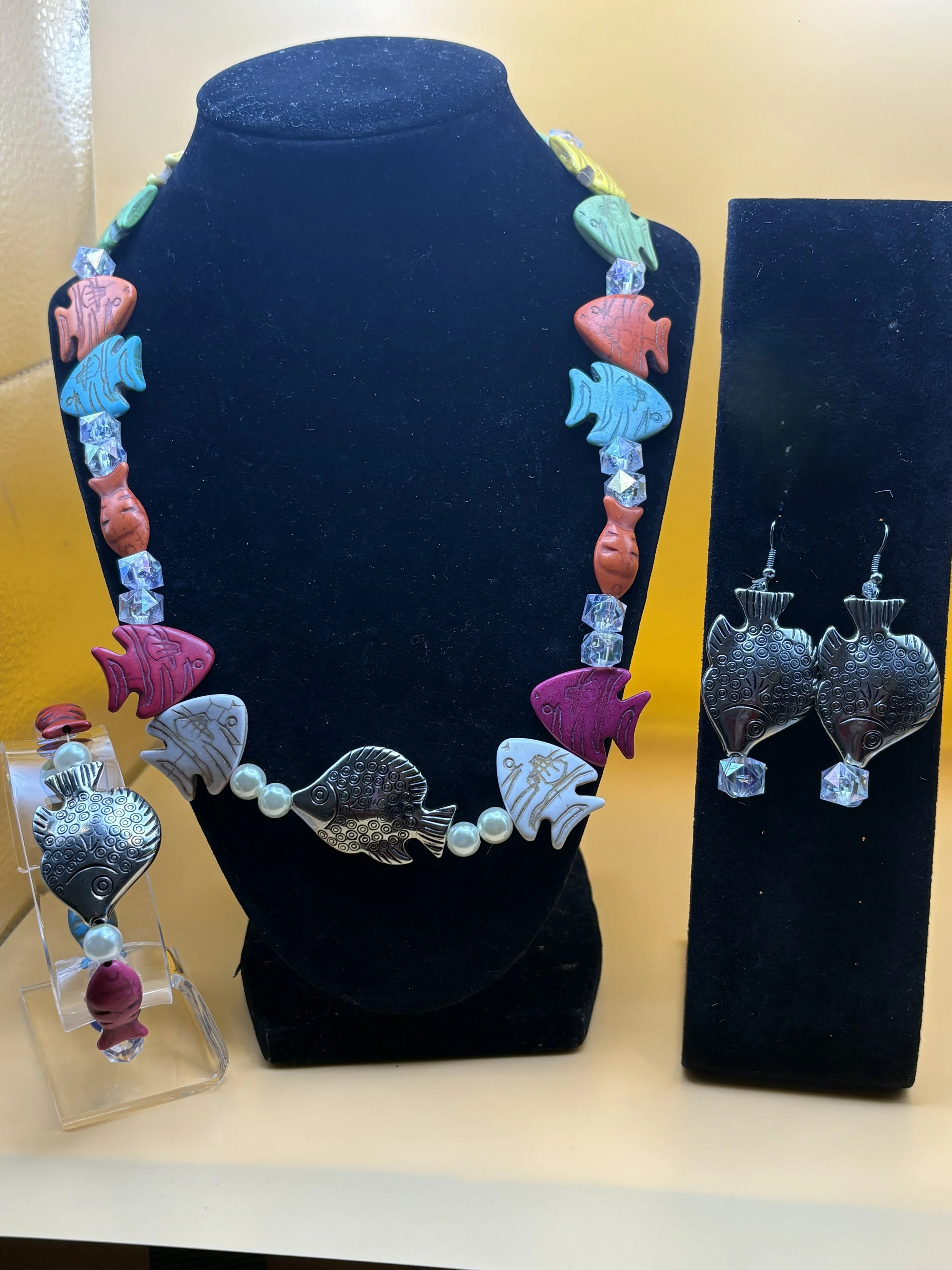 A black jewelry display bust wearing a necklace with fish-shaped beads in various colors and clear crystal-like beads. Next to it is a black jewelry box with matching fish-shaped earrings. To the left, there's a separate stand with additional fish-shaped jewelry, including a bracelet with similar fish beads and a pearl strand.