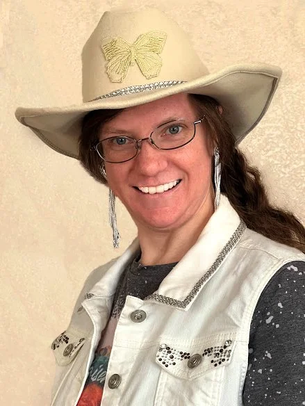Woman wearing a beige cowboy hat with a butterfly decoration, glasses, a white vest with silver embellishments, and a dark shirt with white speckles, smiling at the camera.