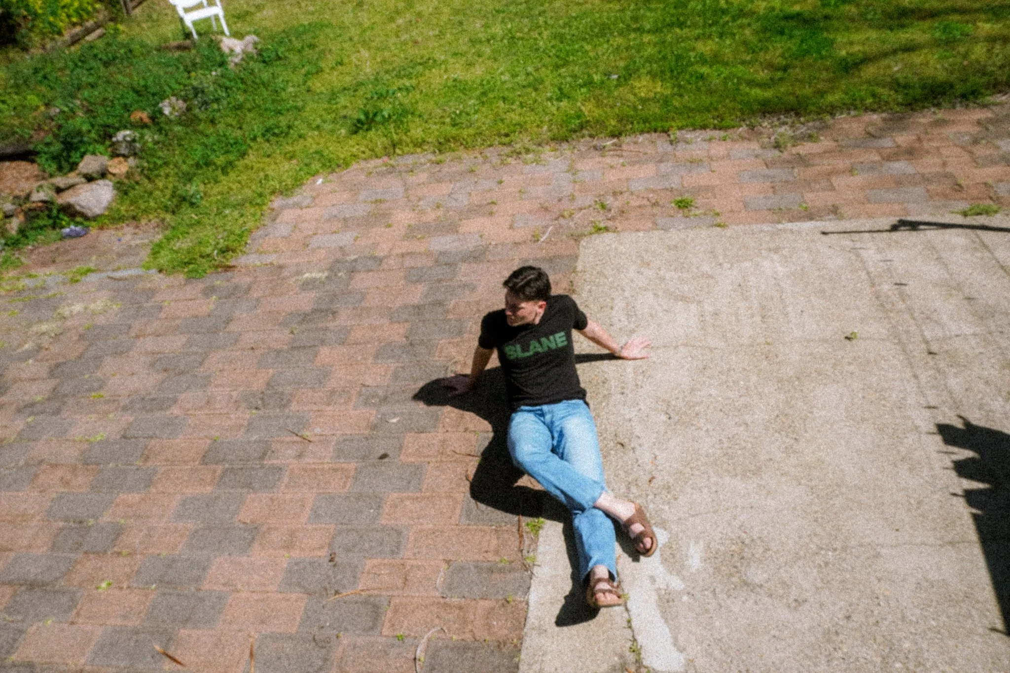 A young person with short dark hair and glasses sitting on a concrete sidewalk, wearing a black T-shirt with green writing, blue jeans, and sandals, at night. There is grass and a garden bed with rocks and plants in the background.