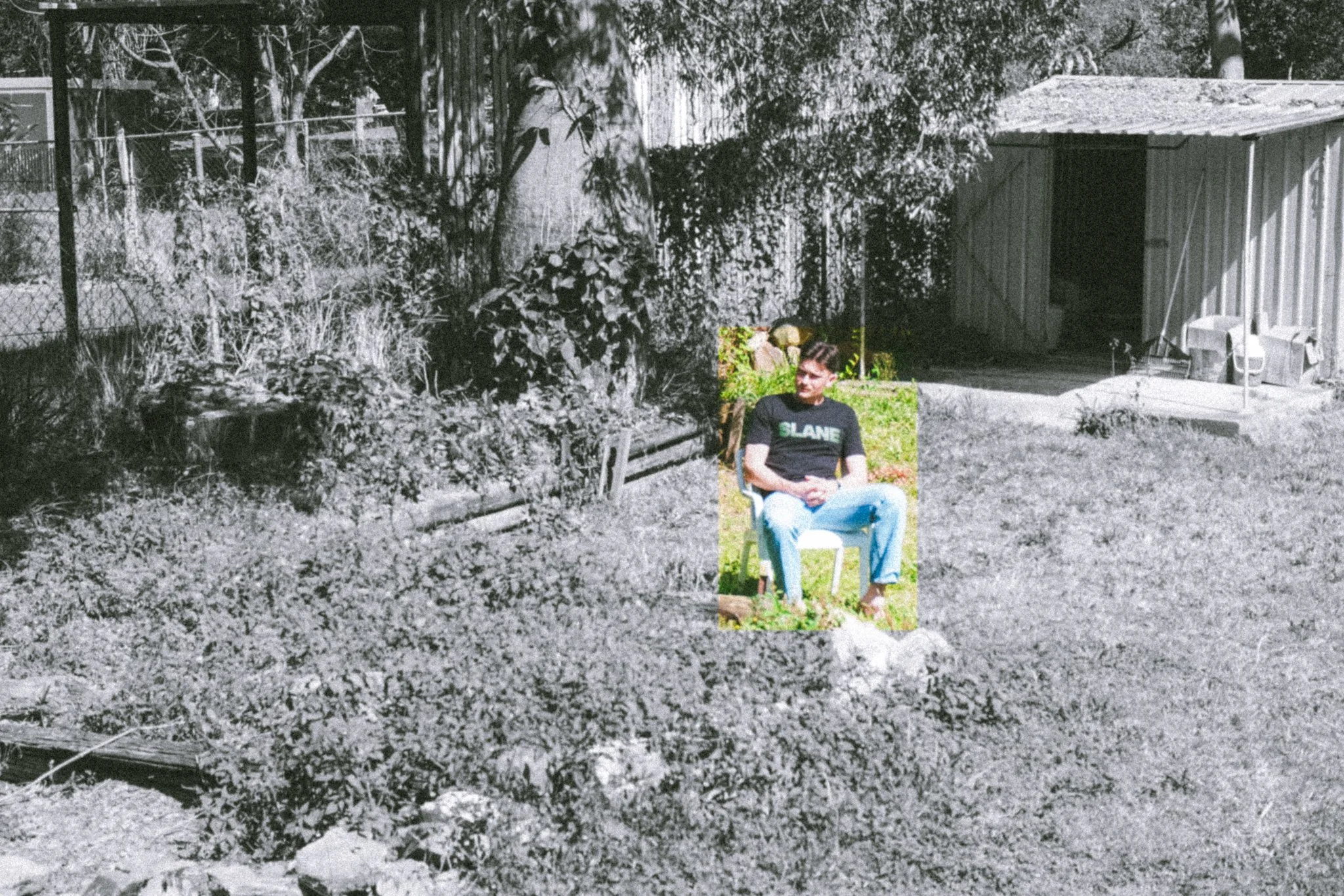 A black and white outdoor scene with a person sitting on a white chair in the grass. The person is wearing a dark T-shirt with the word 'SLANE' on it. There is a small wooden shed and a fence in the background surrounded by trees and bushes.