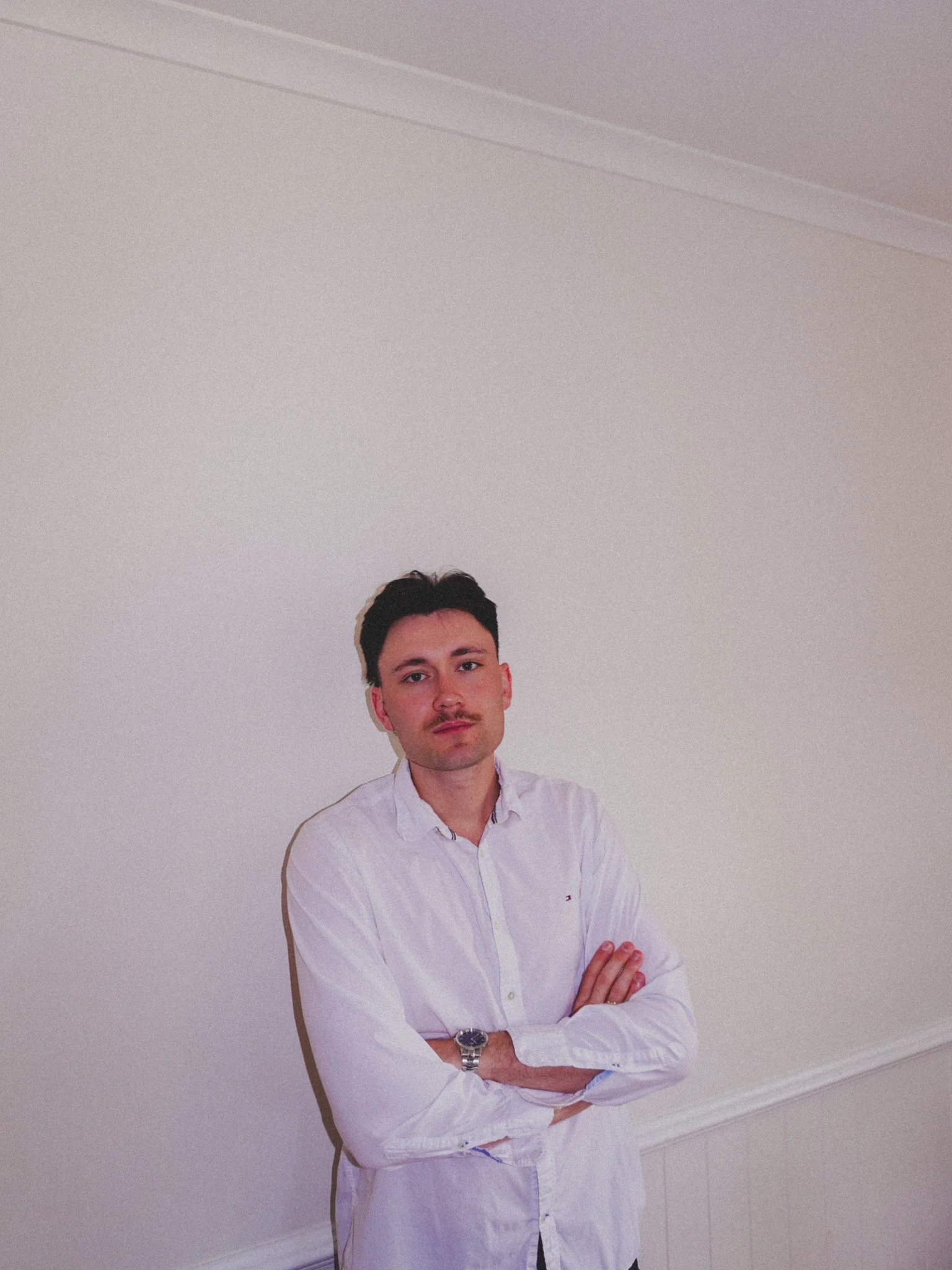 A young man with dark hair, mustache, and light skin, standing against a plain white wall with his arms crossed, wearing a white dress shirt and a watch.