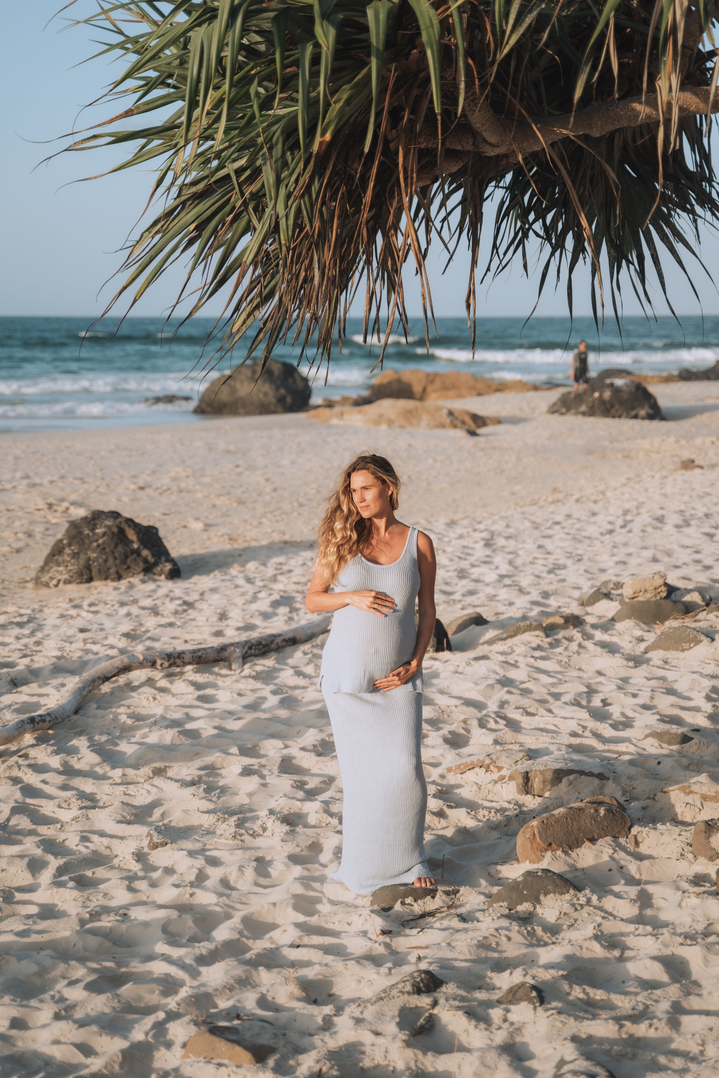 A woman standing on a sandy beach under a thatched roof, with large rocks on the sand and ocean waves in the background.