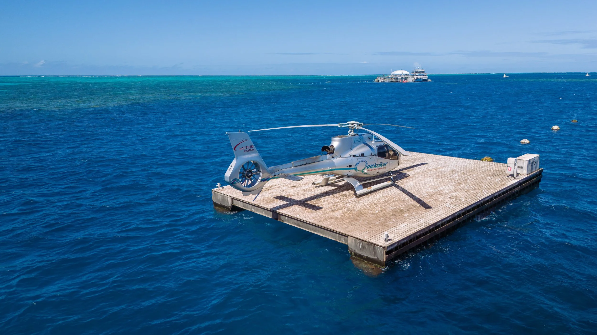A helicopter parked on a floating platform in the ocean with a ship and boats in the background.