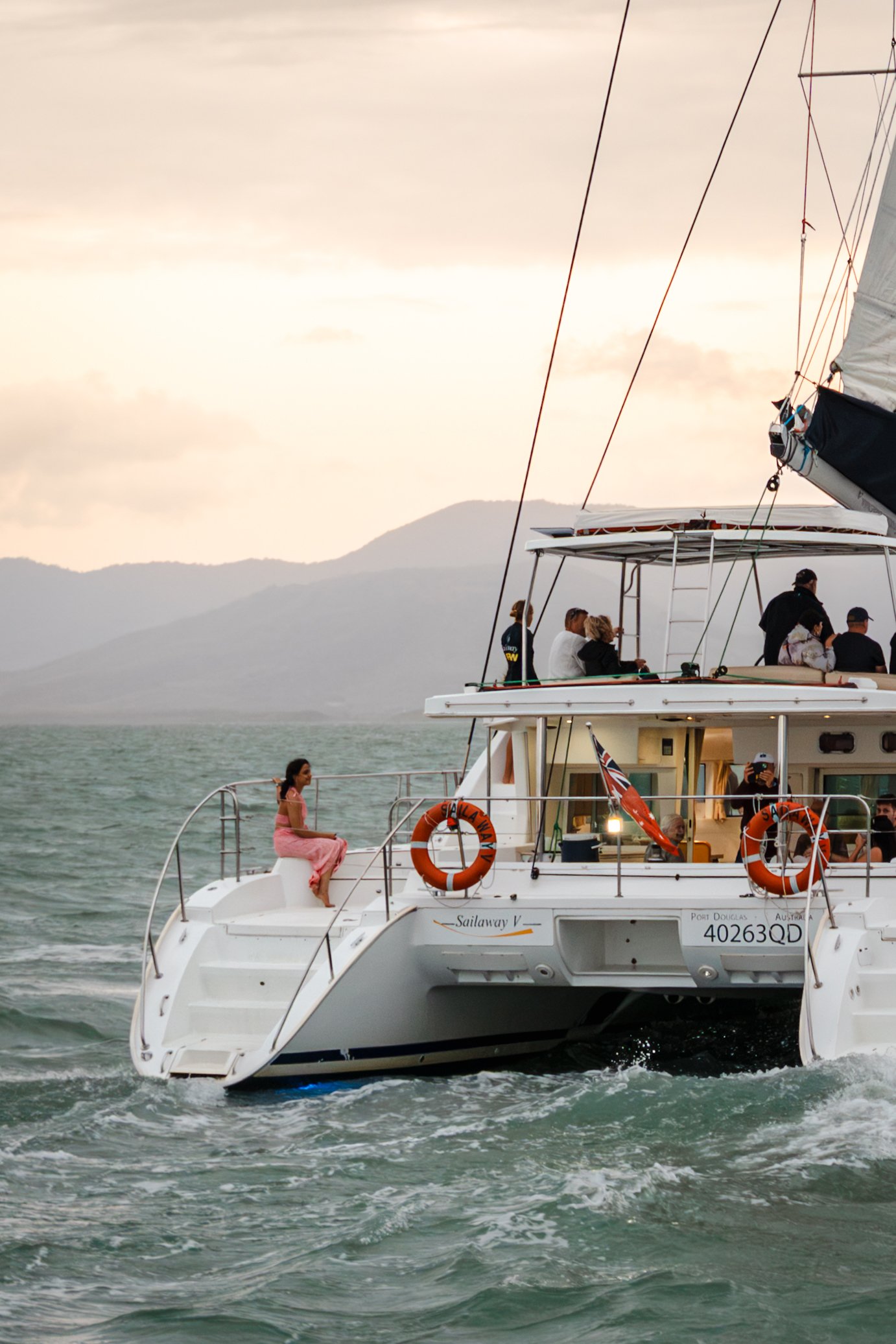 People enjoying a boat trip on a yacht named Sailaway V on the water with a mountain range visible in the background during sunset or sunrise.