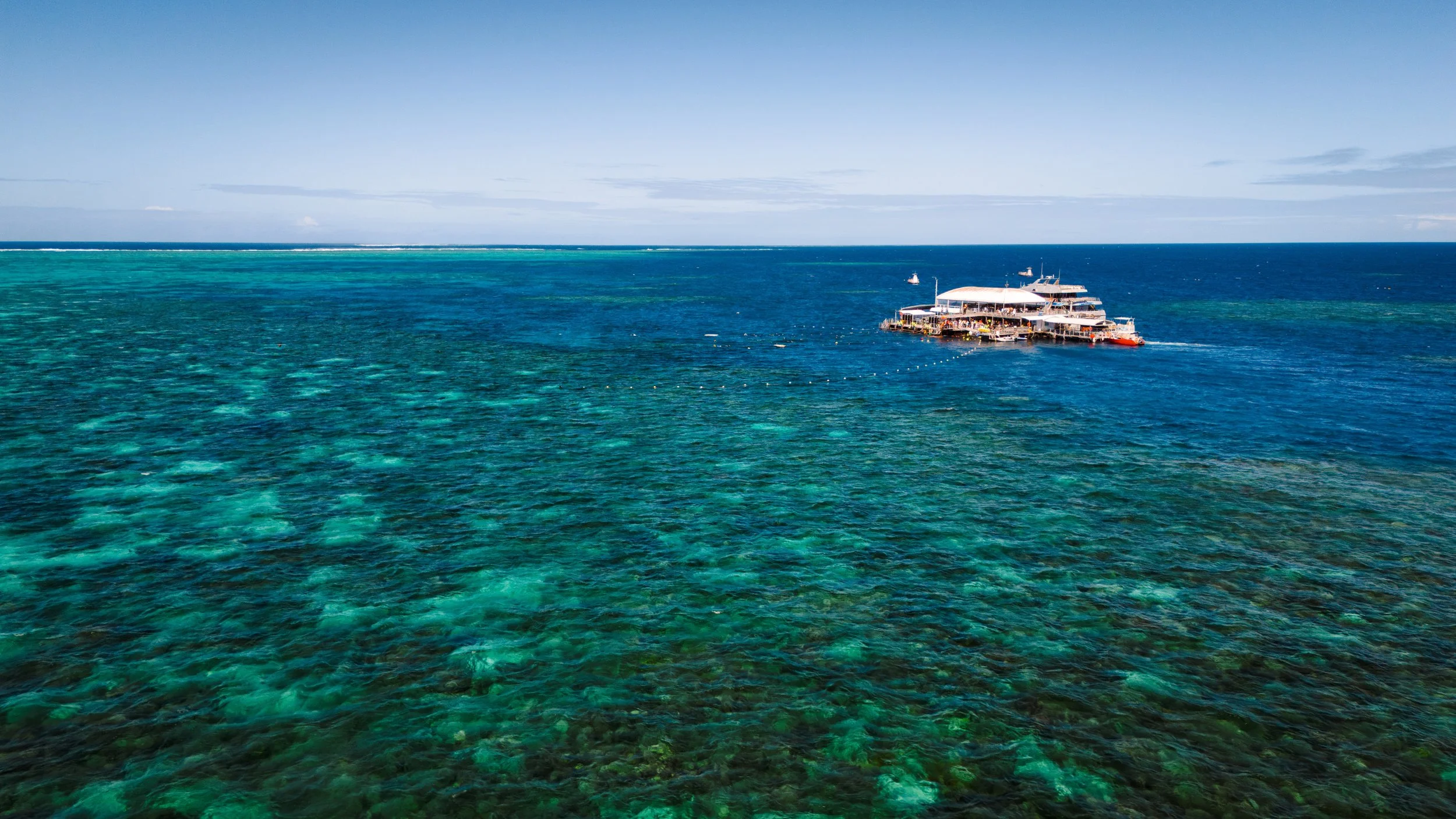 A boat with multiple decks floating on clear blue-green ocean water under a partly cloudy sky.