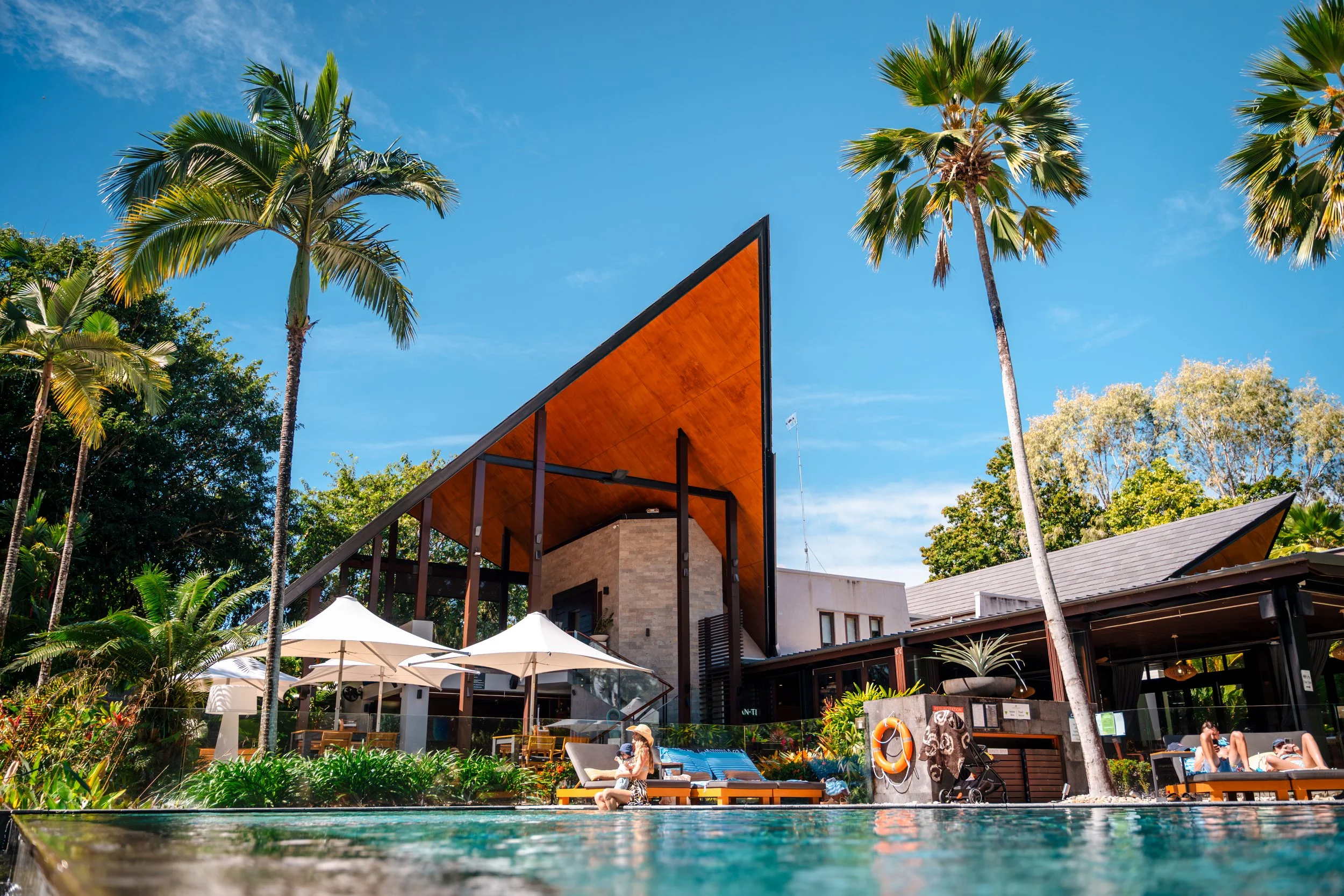 Poolside scene at a modern resort with palm trees, umbrellas, lounge chairs, and people relaxing.