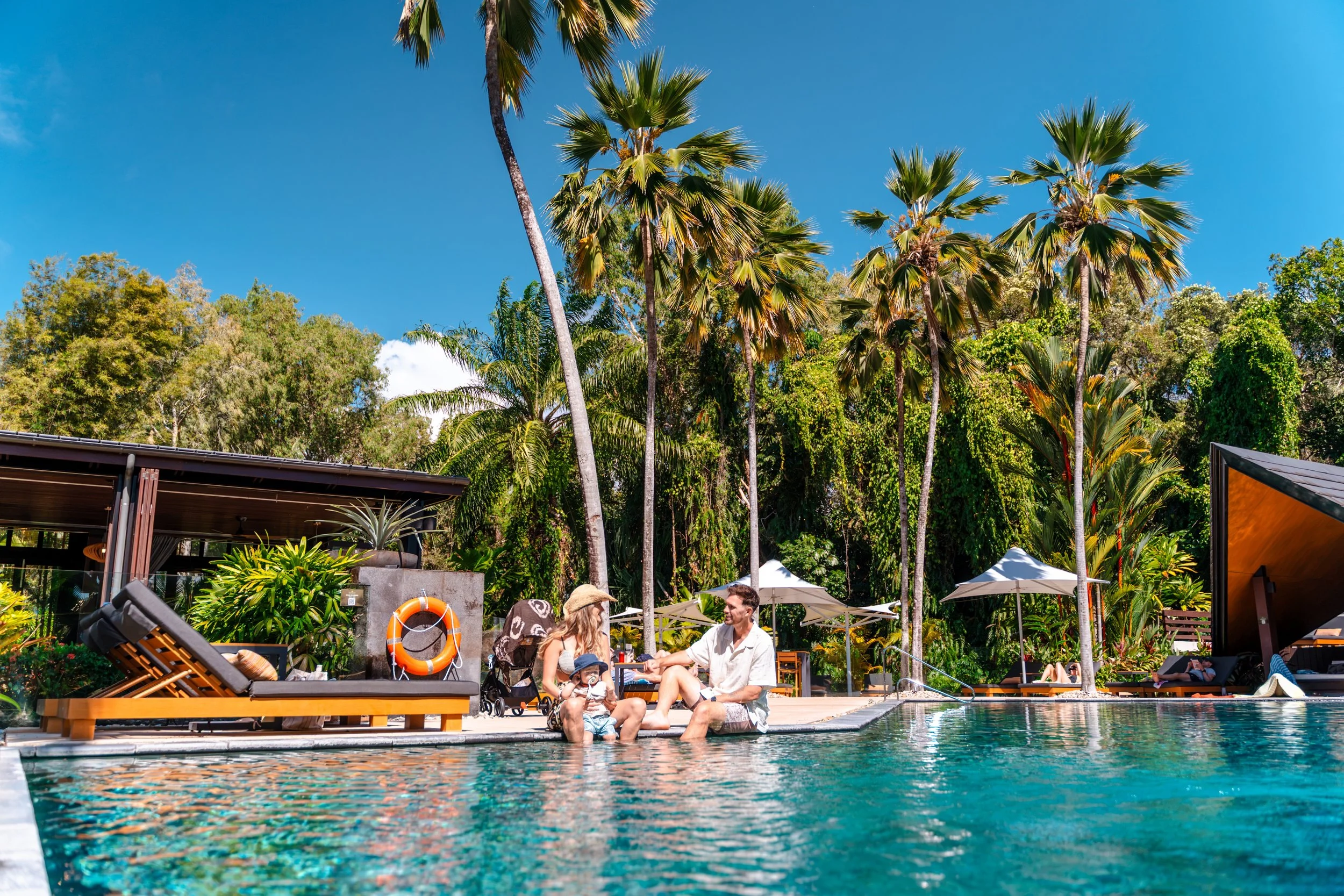 People sitting by a swimming pool surrounded by palm trees and lush greenery under a clear blue sky.