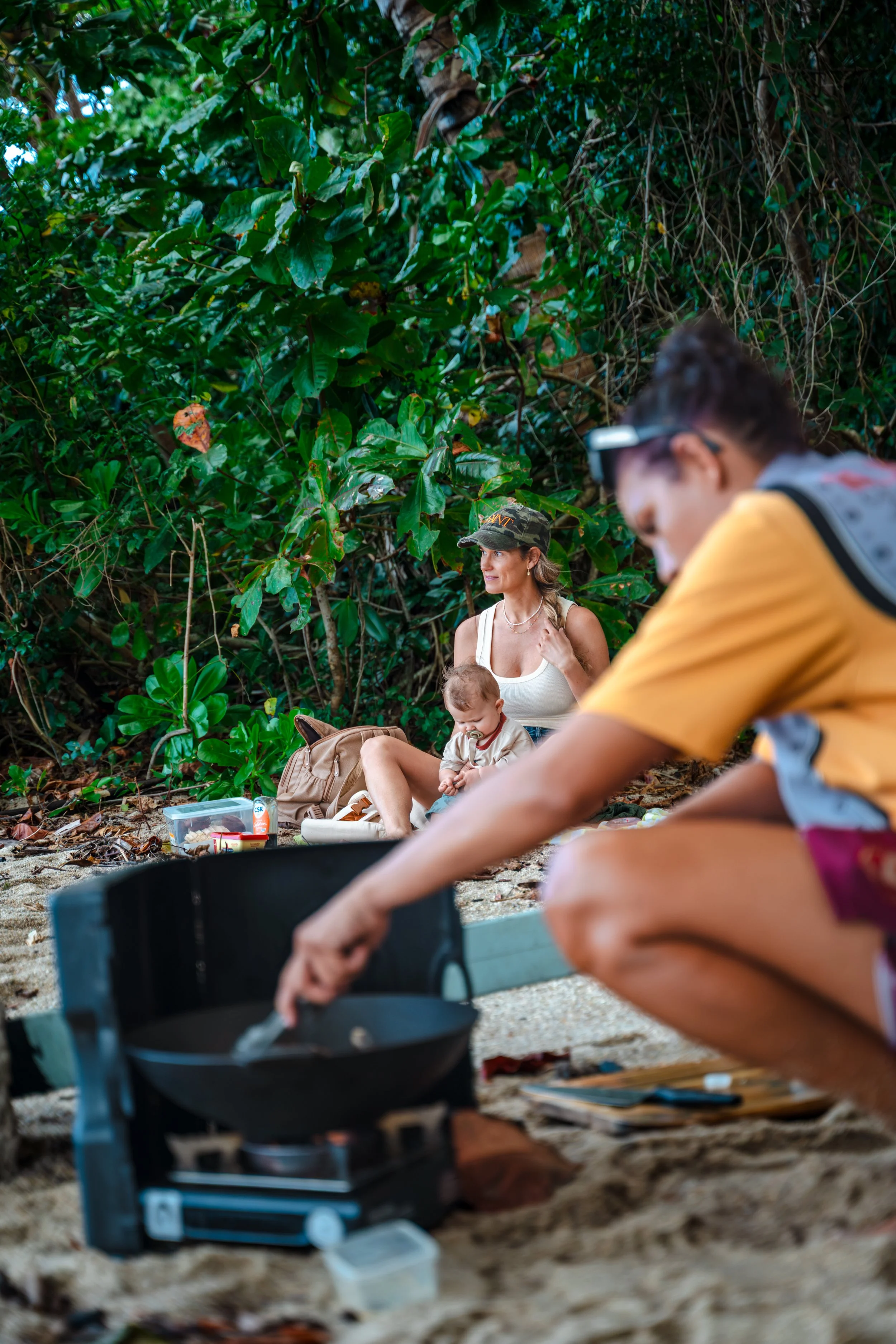 People sitting on the ground in a jungle area, with a woman holding a baby, and another person cooking on a portable stove.