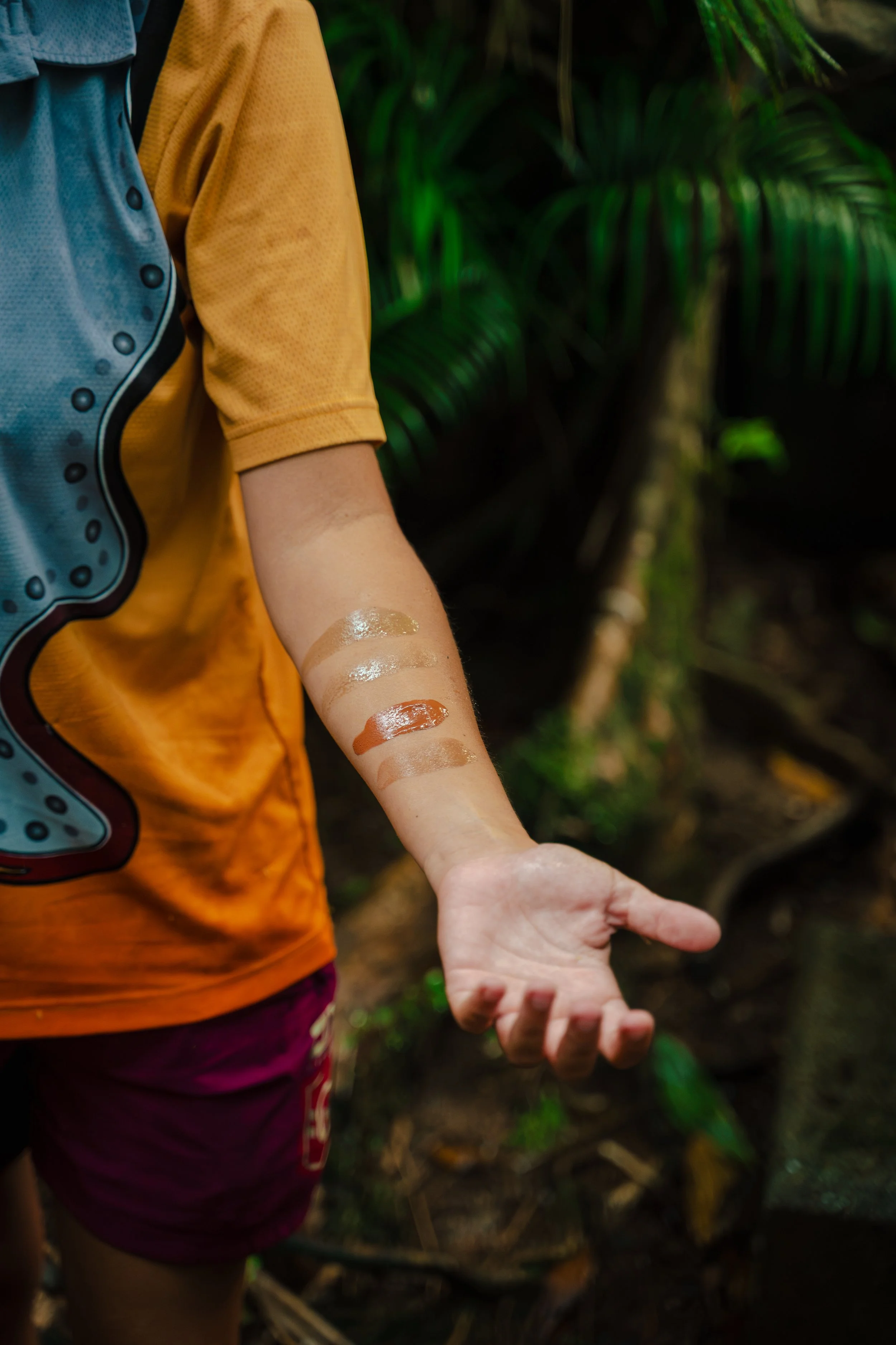 Close-up of a child's arm with swatches of different colored paints, wearing a yellow shirt and maroon shorts, standing outdoors with green tropical plants in the background.