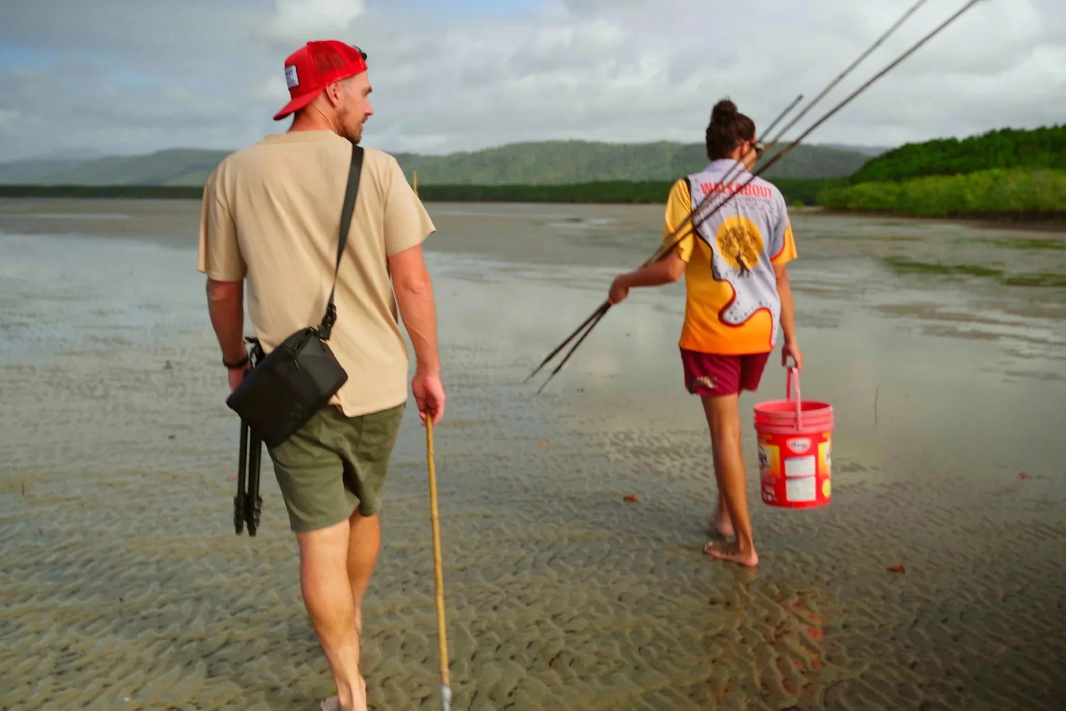 Two people walking at the beach with fishing poles, one person with a red bucket, overcast sky with hills in the background.
