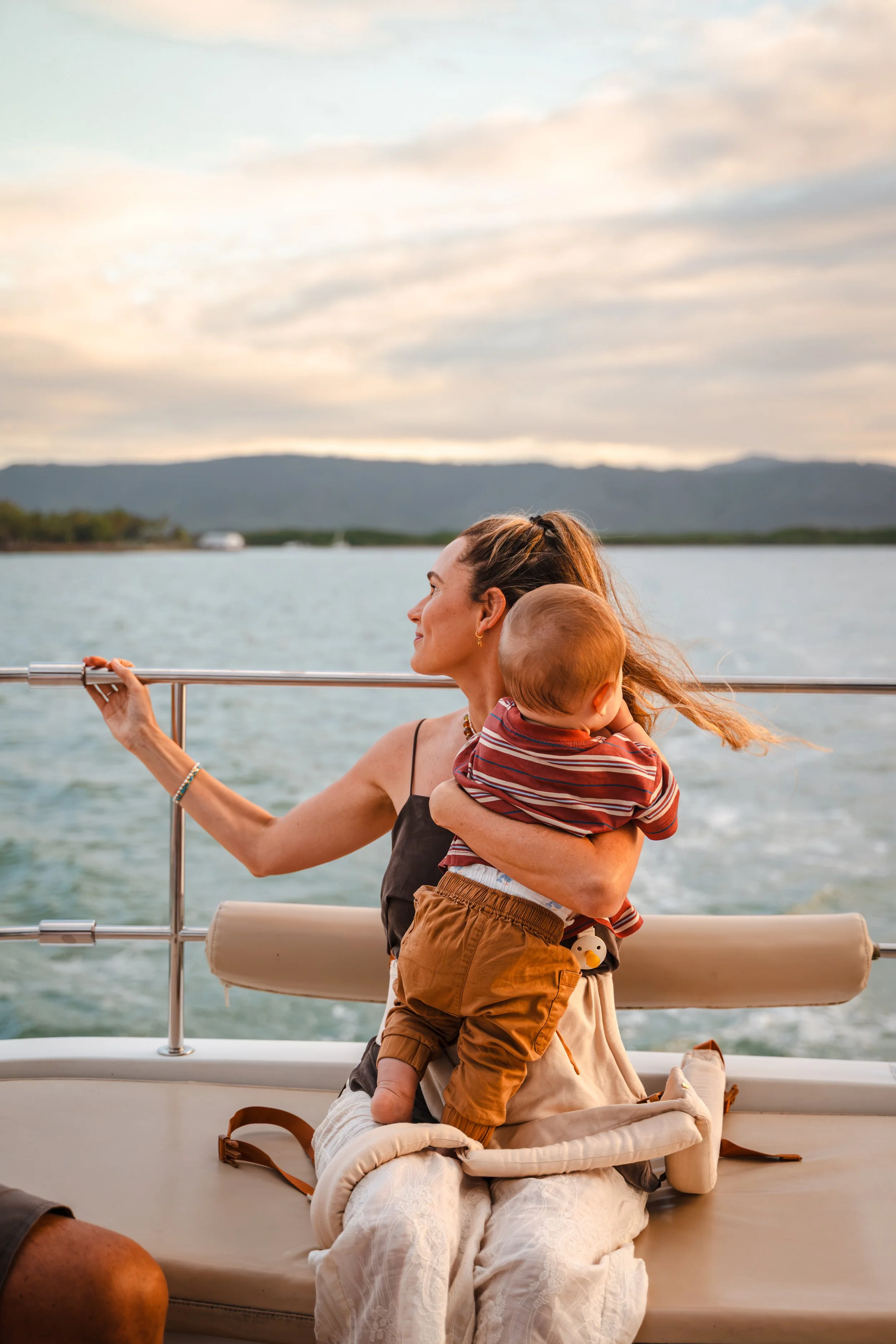 A woman holding a toddler on a boat with water and mountains in the background during sunset.