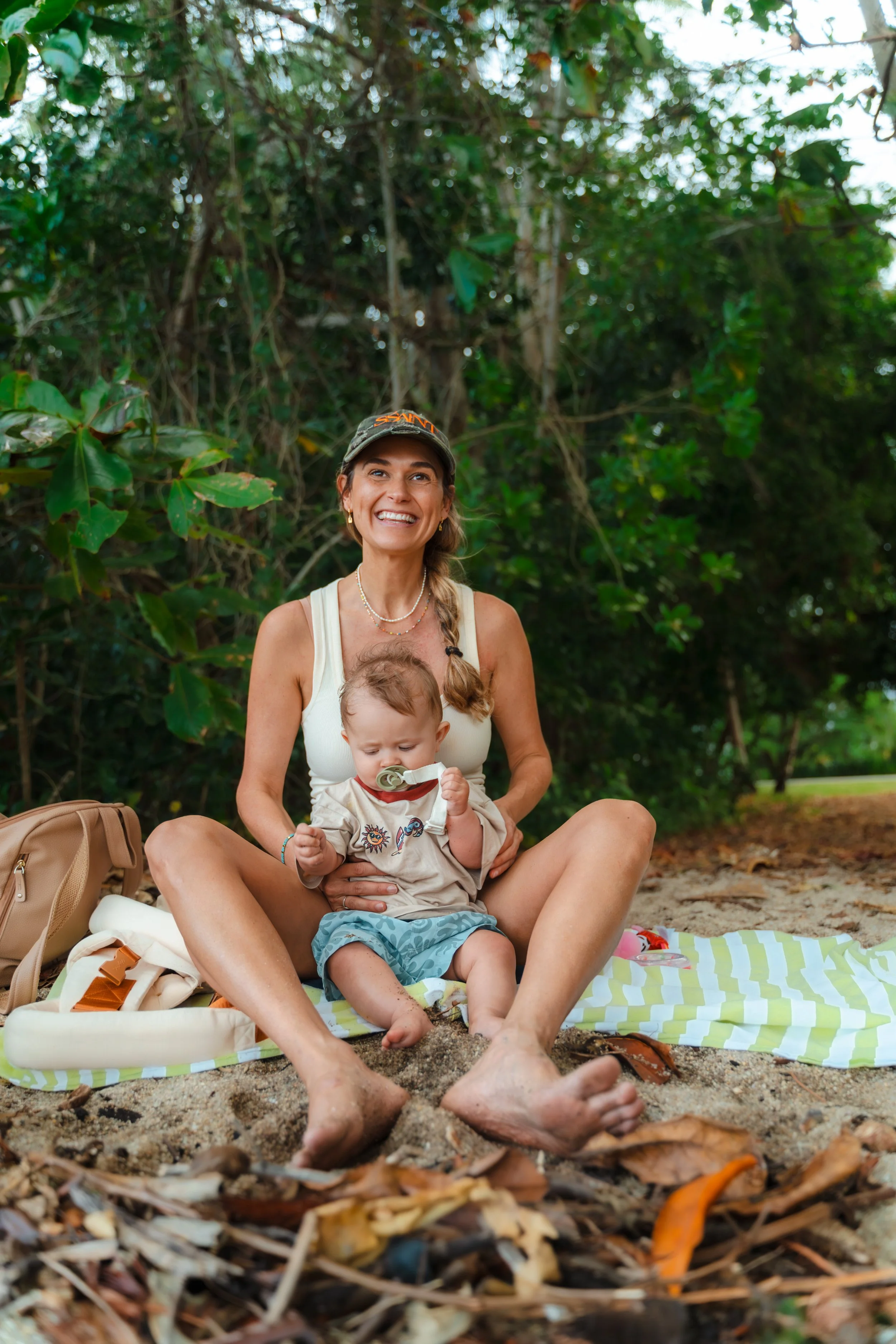 A woman smiling and sitting cross-legged on a beach towel with a young child sitting on her lap, surrounded by nature in a forested outdoor setting.