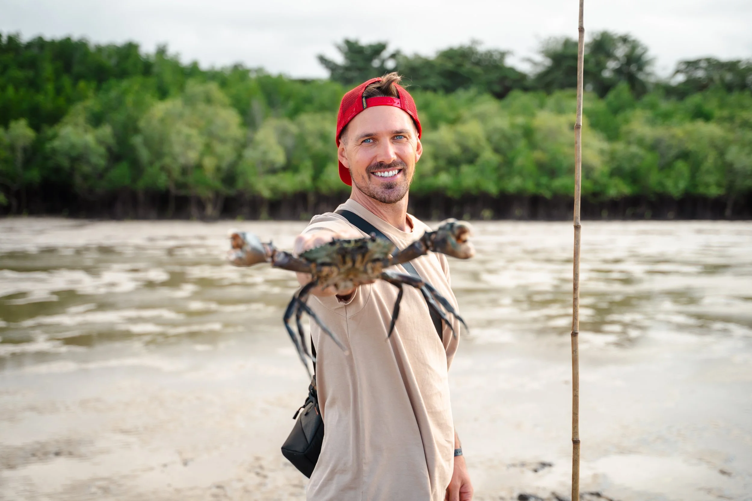A smiling man with a red cap backwards, holding a crab in his hand, standing on a beach with lush greenery and water in the background.