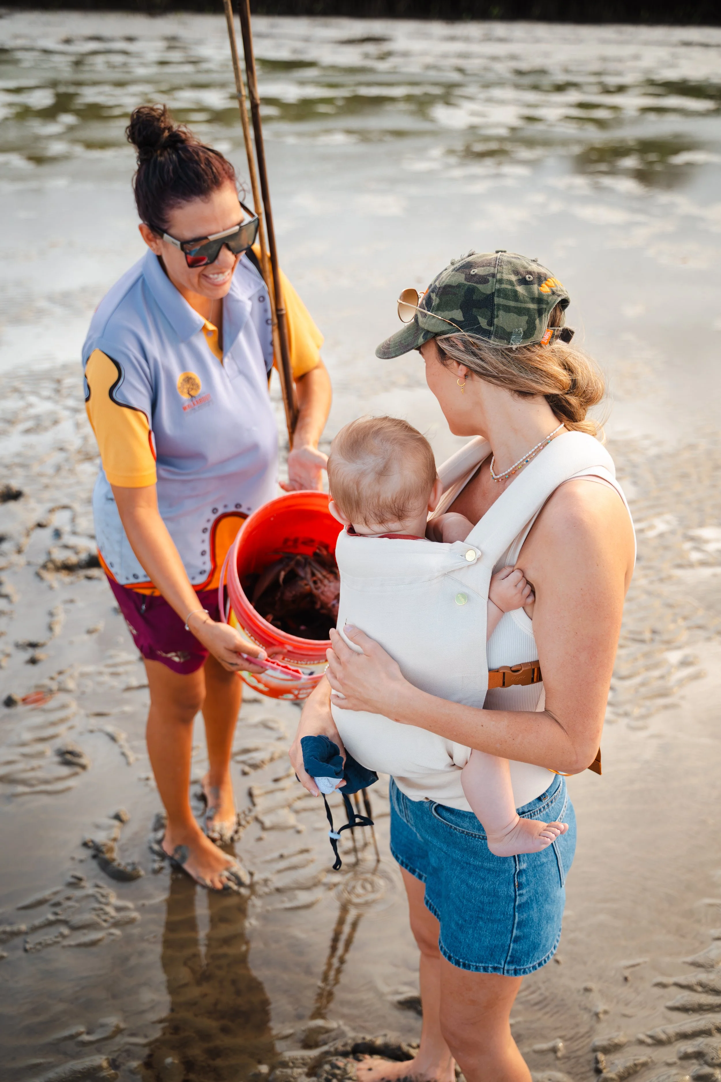 Woman holding a child in a baby carrier on a beach, while another woman offers a bucket of what appears to be a large marine animal, possibly a stingray, on the shoreline.