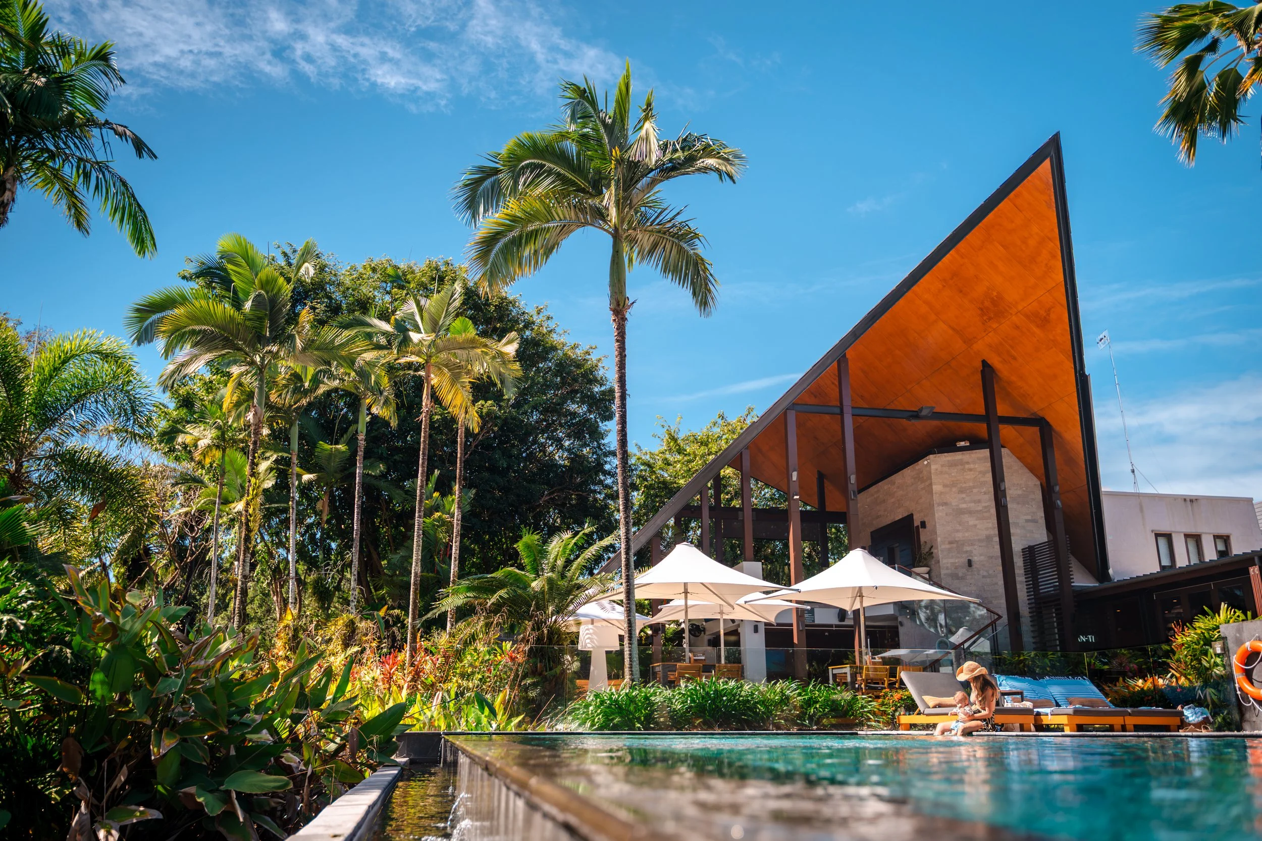Resort pool surrounded by lush tropical trees, with a woman and child sitting on the poolside, overlooking white umbrellas and modern buildings with a wooden roof and palm trees in the background under a bright blue sky.