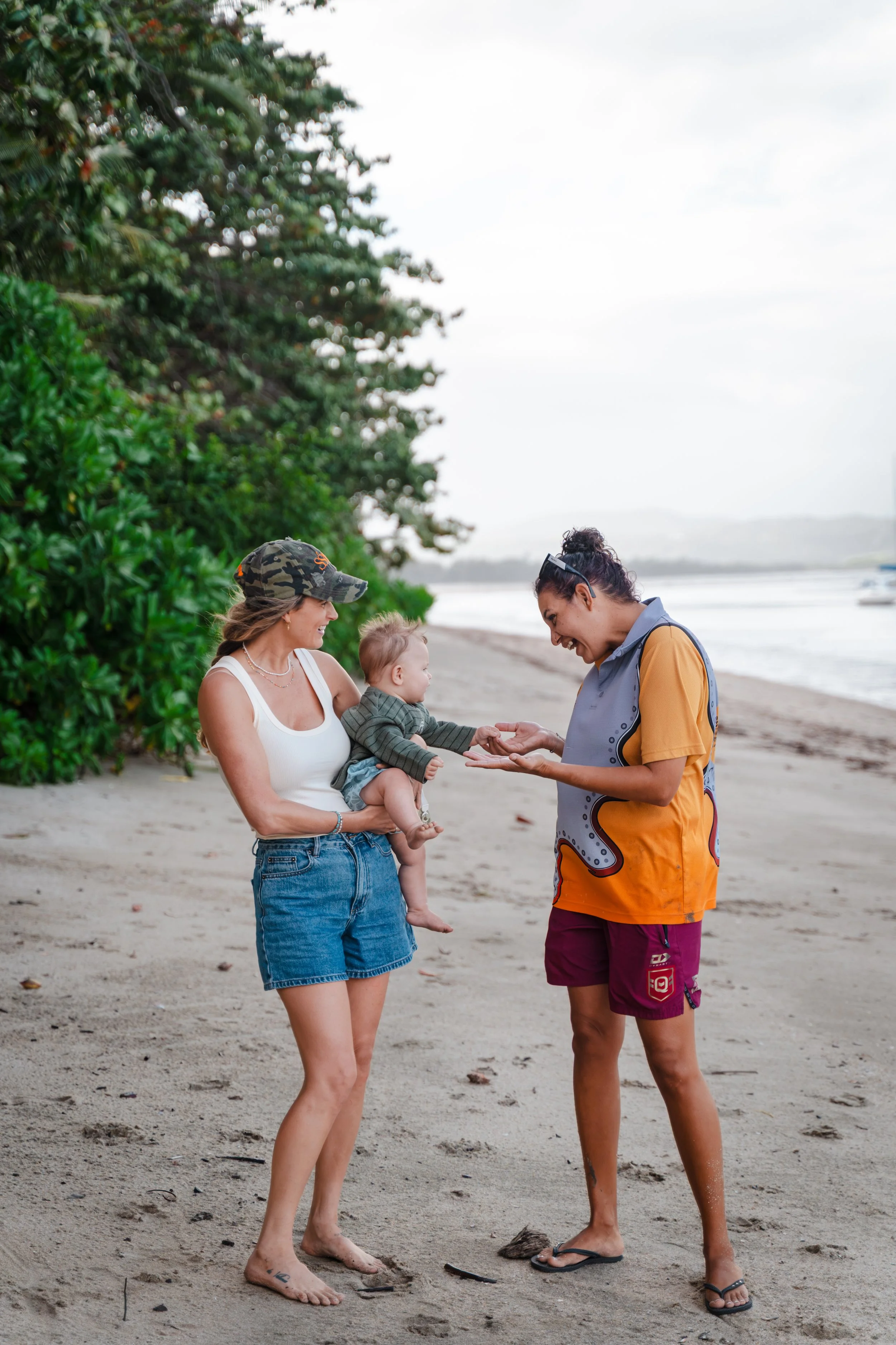 Two women and a baby standing on a sandy beach near water, with greenery on the left. One woman wearing a white tank top, denim skirt, and camouflage cap is holding the baby. The other woman, dressed in orange and purple athletic wear, is showing som