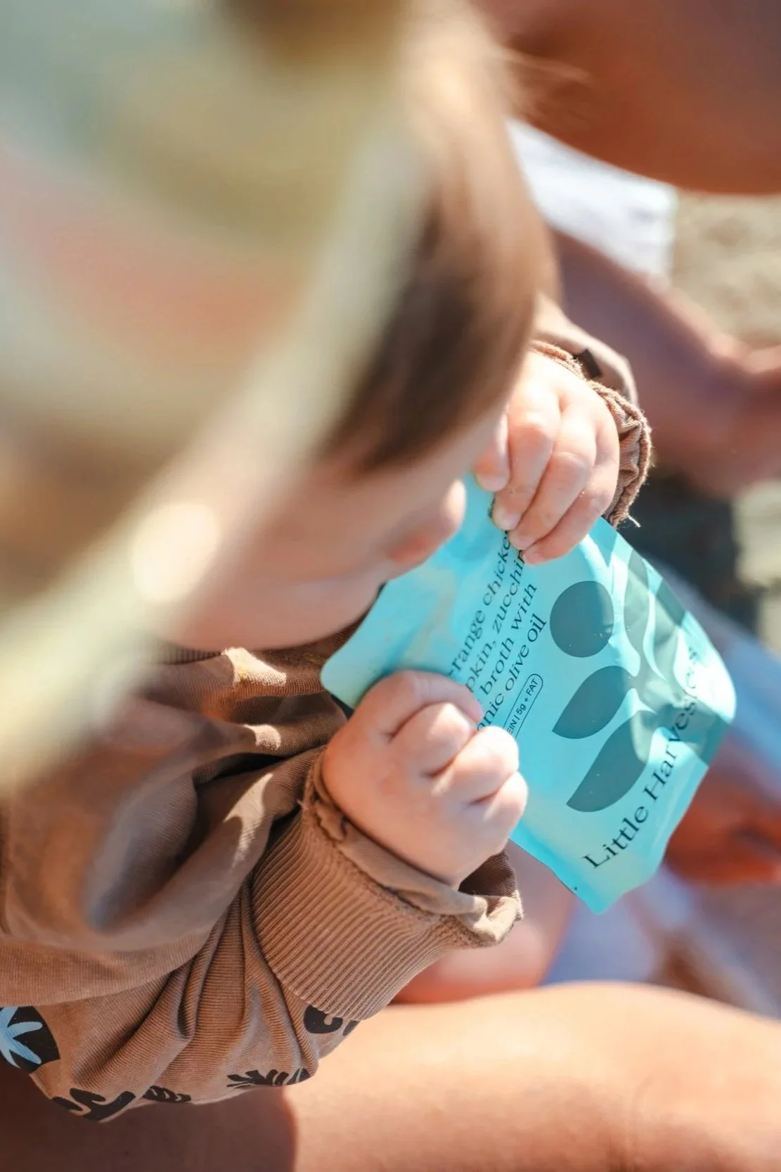 A young child holding a blue packet of organic chicken with tea. The child is wearing a brown long-sleeve shirt with black patterns and has light skin and reddish hair. The focus is on the child's hand gripping the packet, with the child's face blurred in the background.
