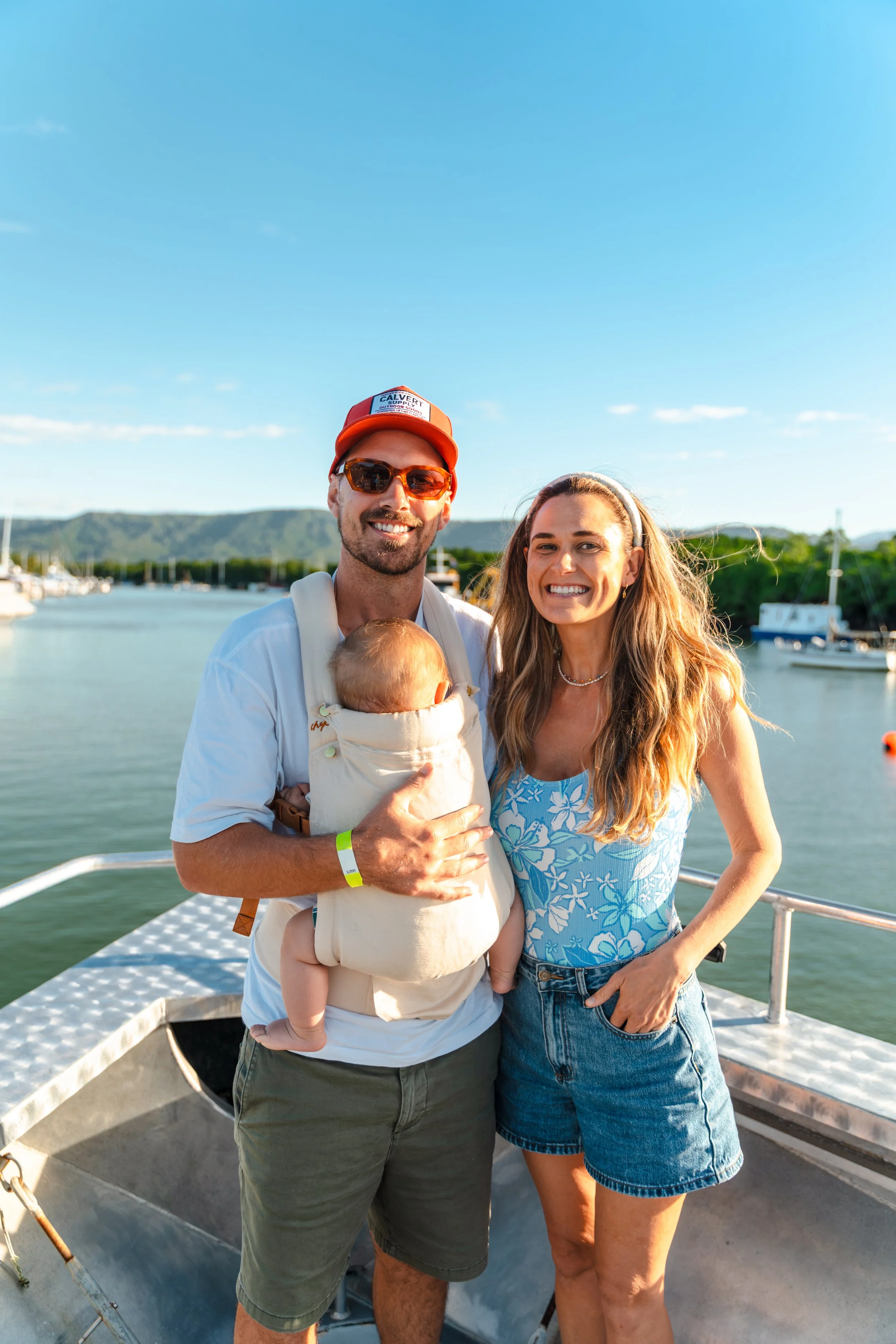 A smiling man and woman standing on a boat by a harbor, with mountains and boats in the background, during daytime.