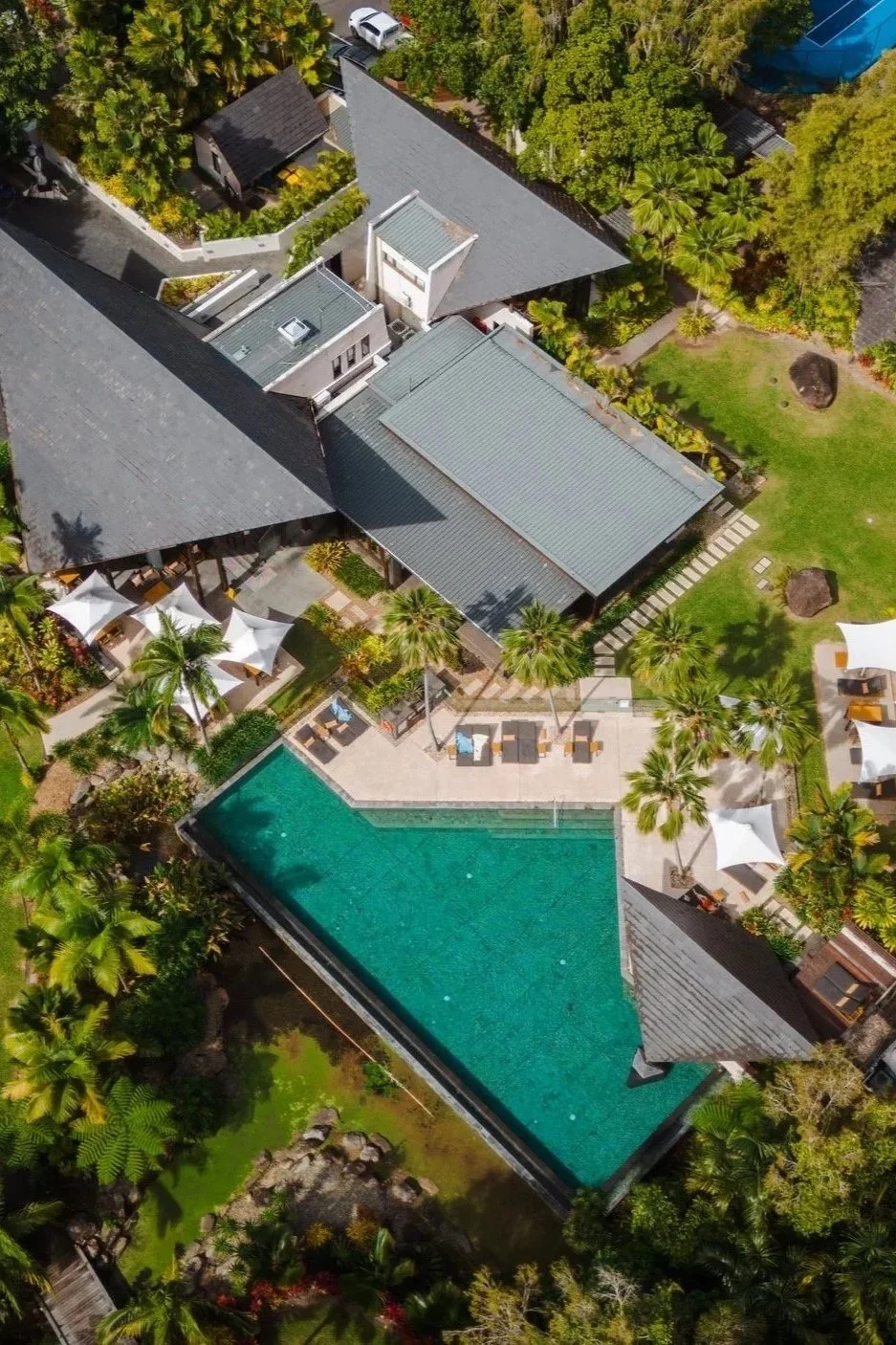 Aerial view of a luxury resort featuring a large swimming pool, surrounded by lounging chairs, umbrellas, palm trees, and lush greenery. There are buildings with grey rooftops and patios, along with landscaped gardens and walking paths.