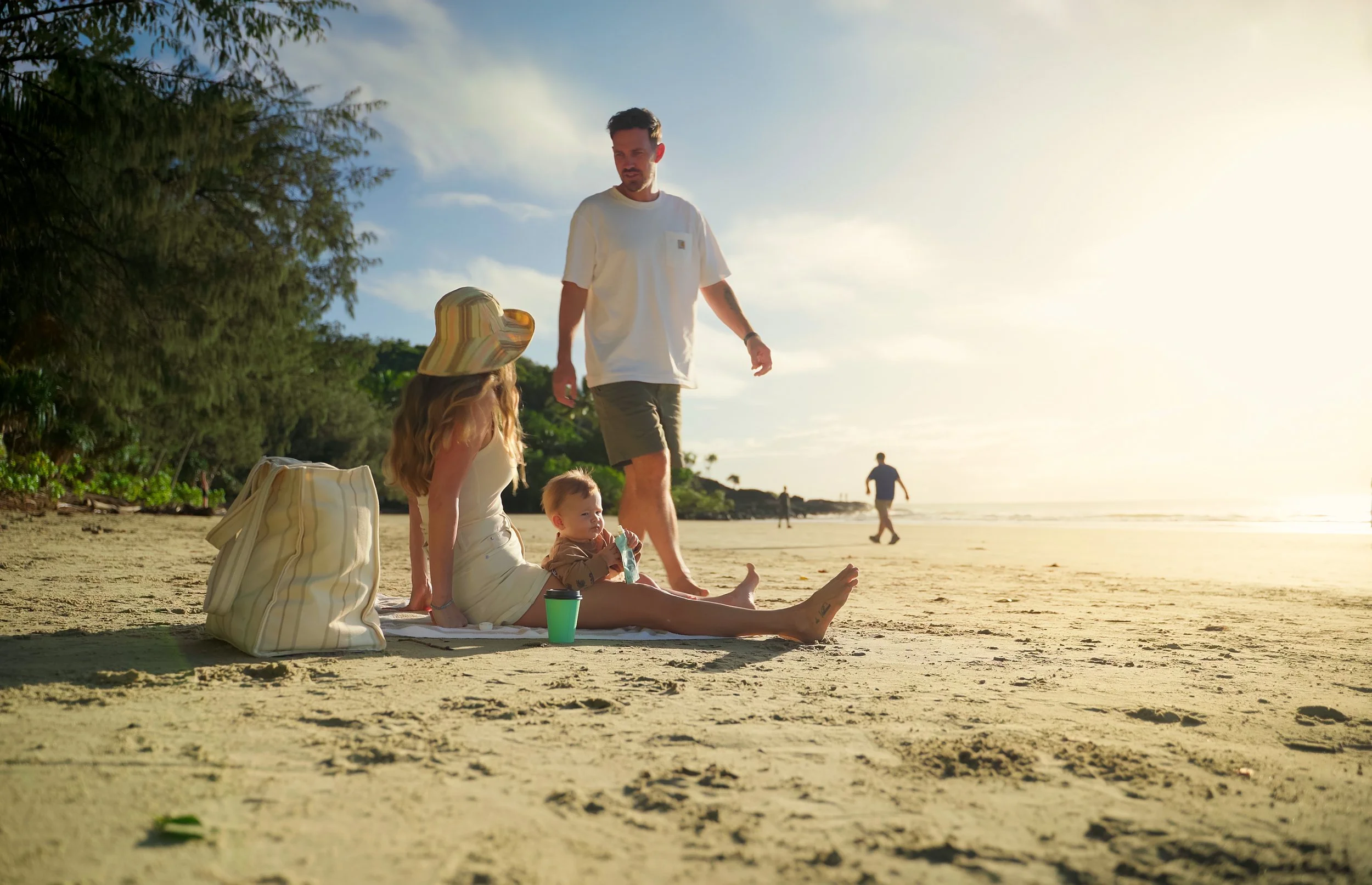A family relaxing on a sandy beach during sunset. A woman sits on the sand with a child and a dog with a striped hat nearby. A man stands looking at the family, with another person walking in the background.