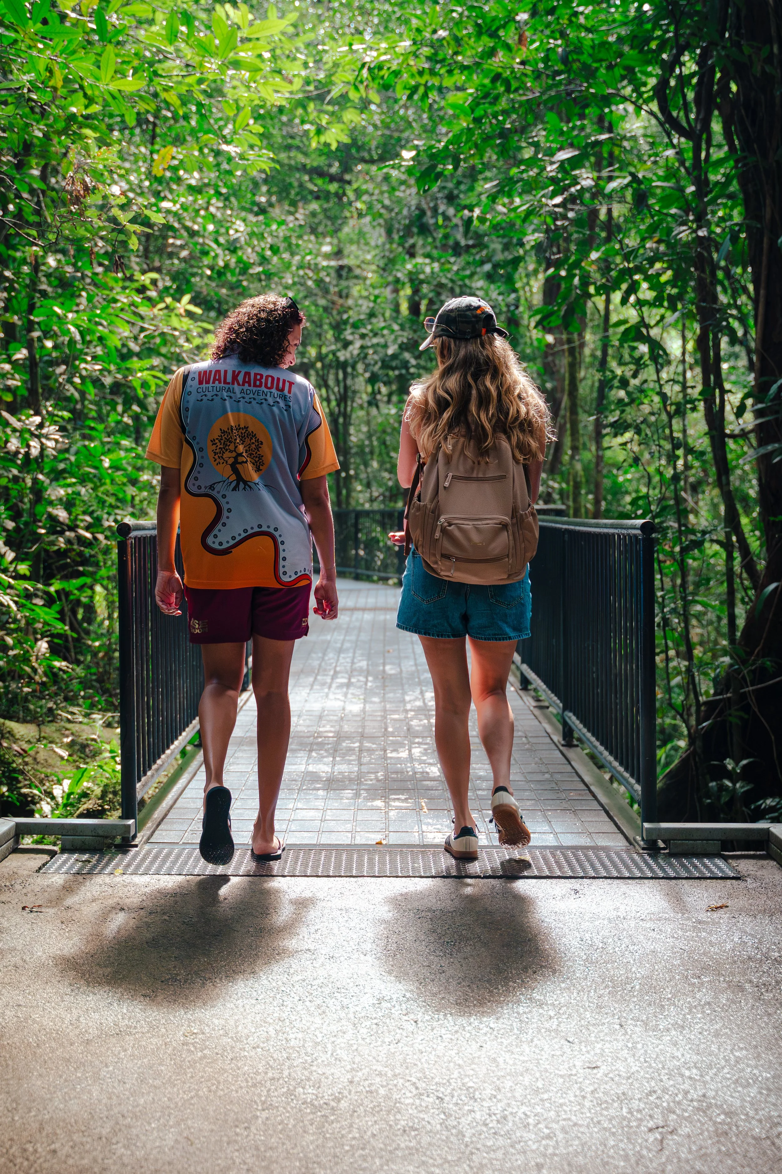 Two women walking on a brick pathway through a lush green forest with trees and sunlight filtering through the leaves.