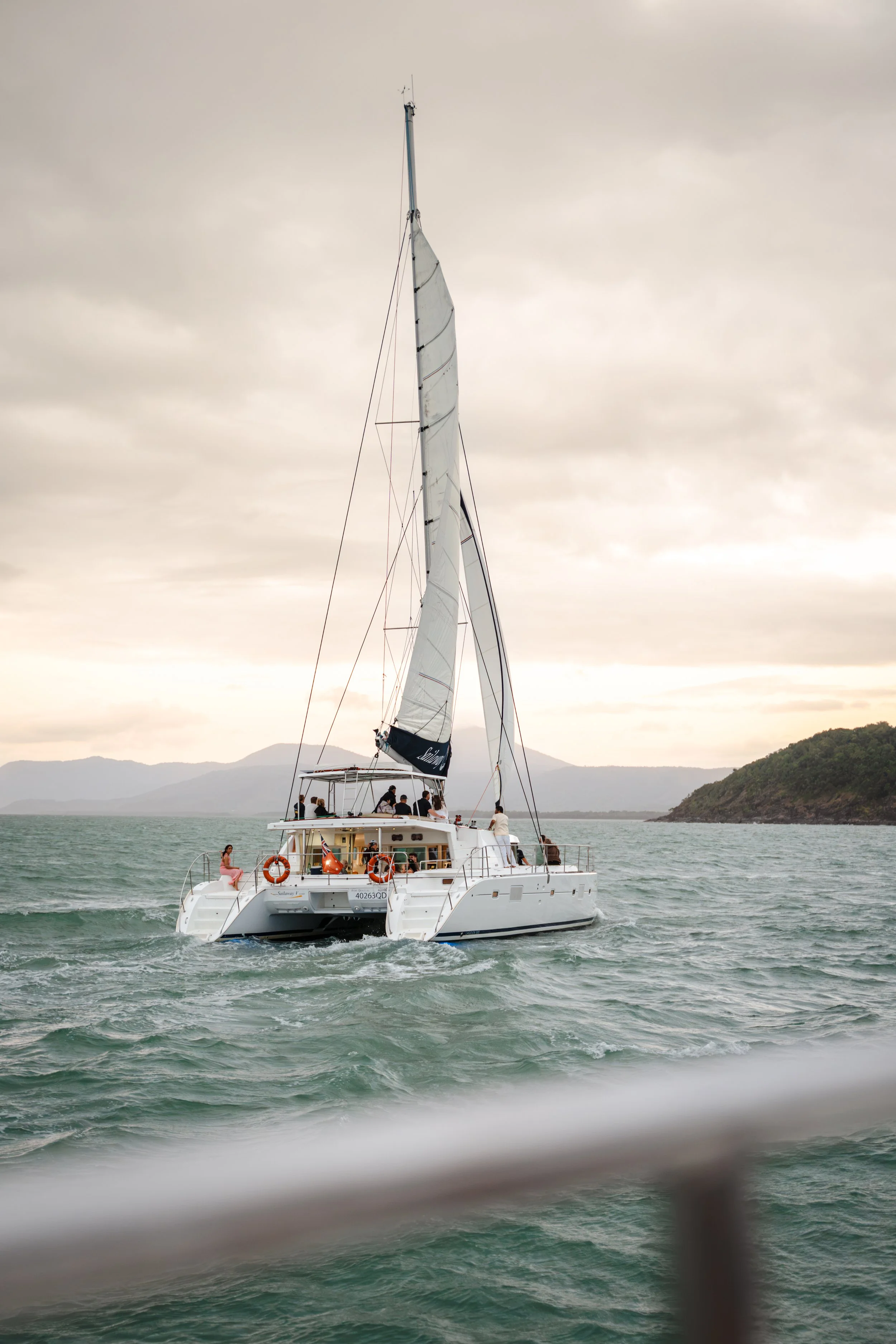A group of people enjoying a boat ride on a white sailboat with a large sail, on a body of water with distant mountains and cloudy sky in the background.