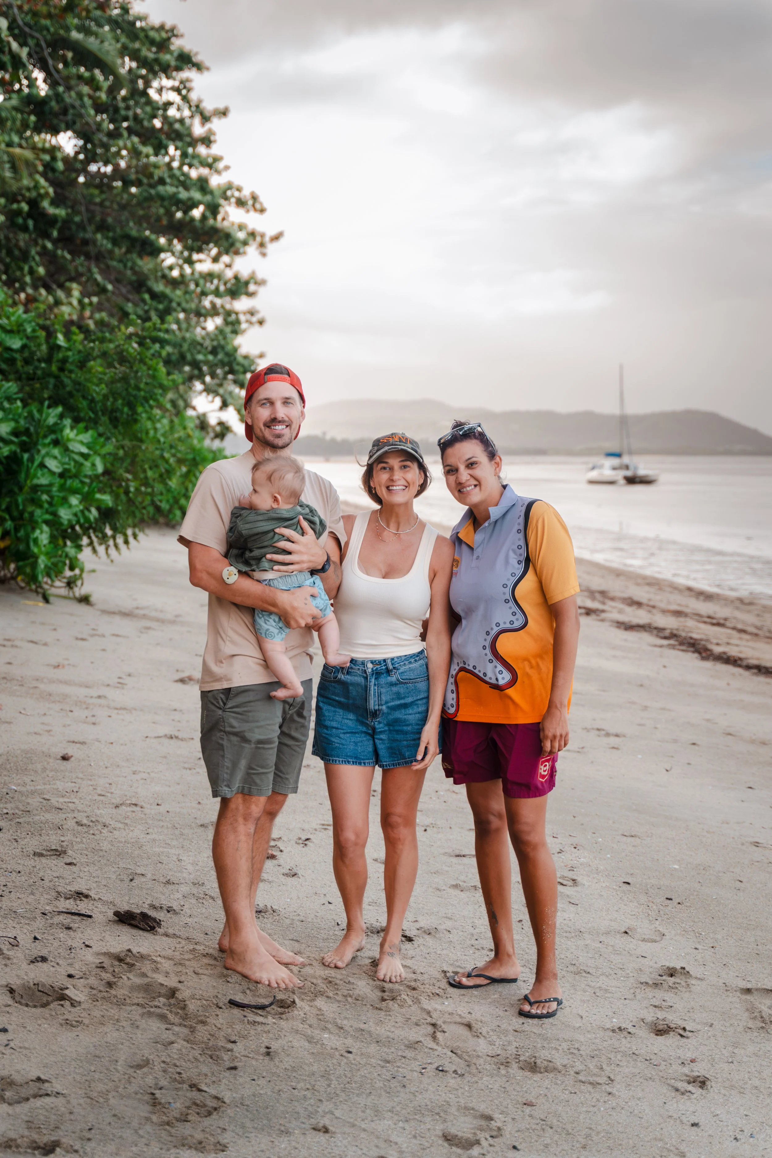 A group of four people, including a man holding a baby, two women, standing on a sandy beach with water and sailboats in the background, and trees on the left side, during overcast weather.