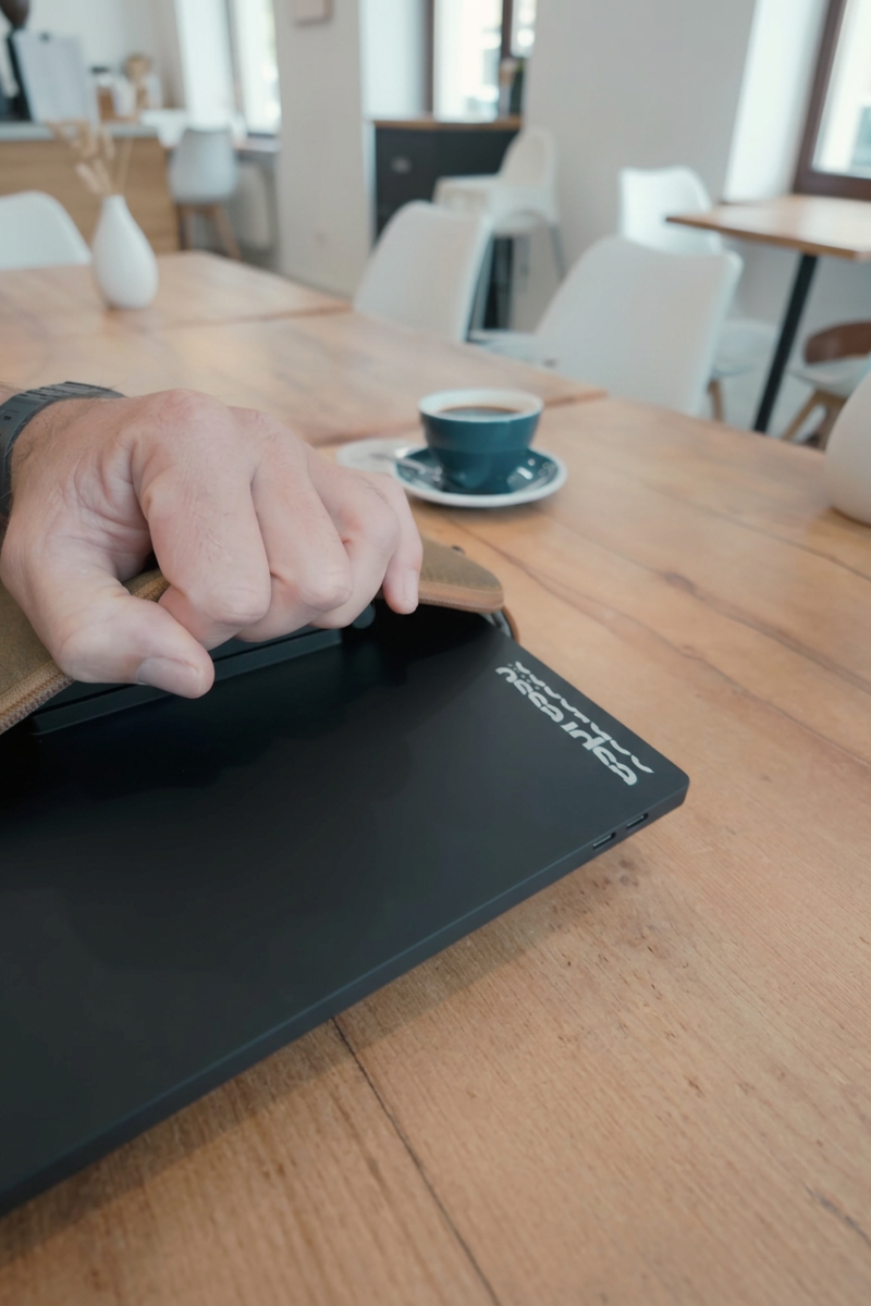 A person opening a portable laptop on a wooden table in a bright cafe with white chairs, a cup of coffee, and white vases as decor.