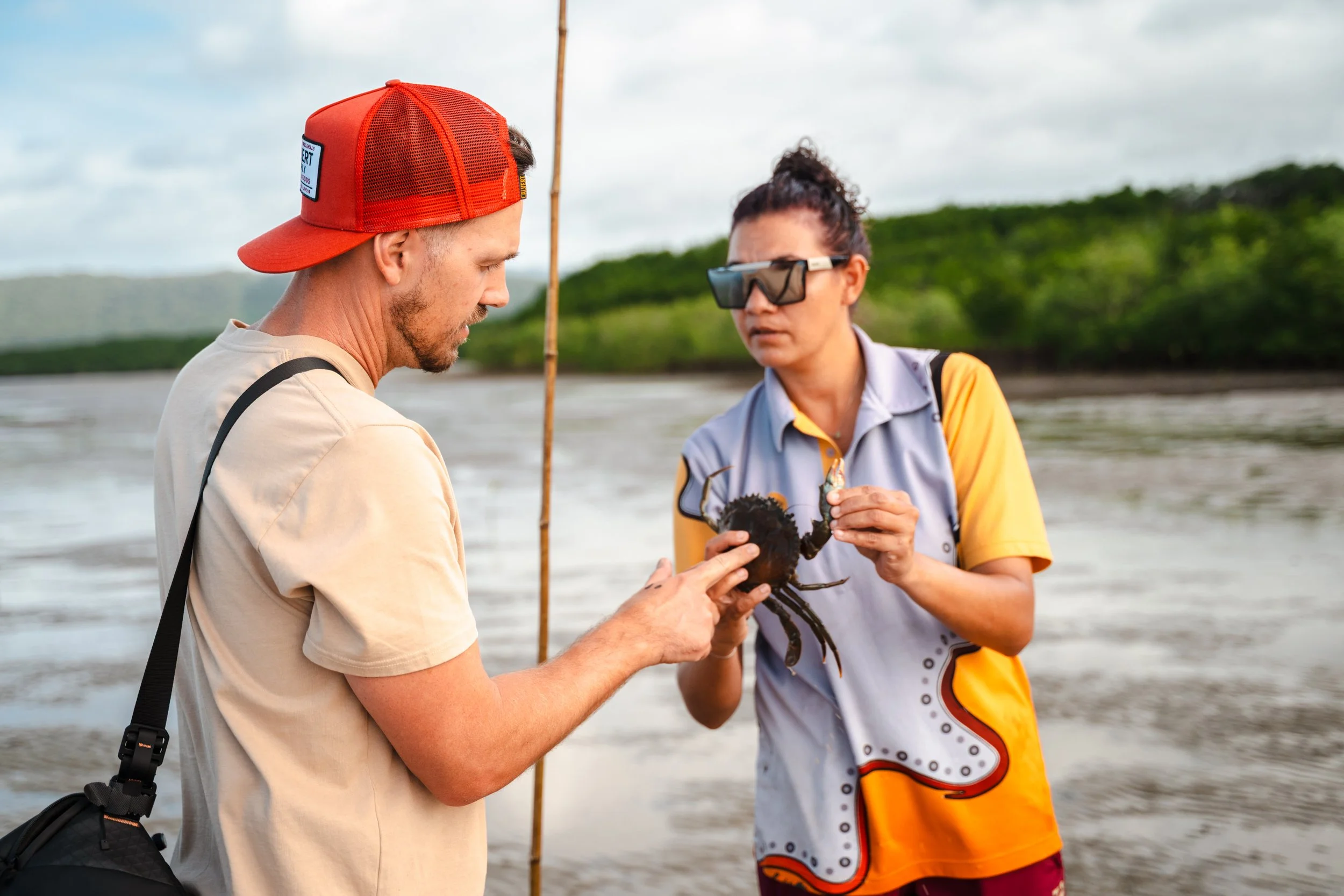 Two people on a beach, one holding a lobster and the other examining it, with water and greenery in the background.
